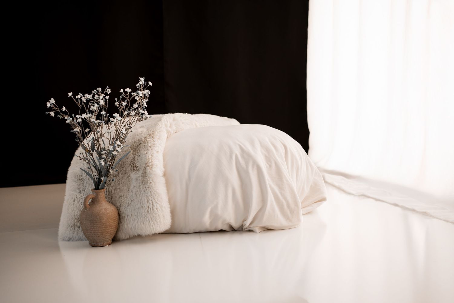 Cozy white bedding and a faux fur throw on a white floor beside a small terracotta vase with dried flowers against dark curtains.