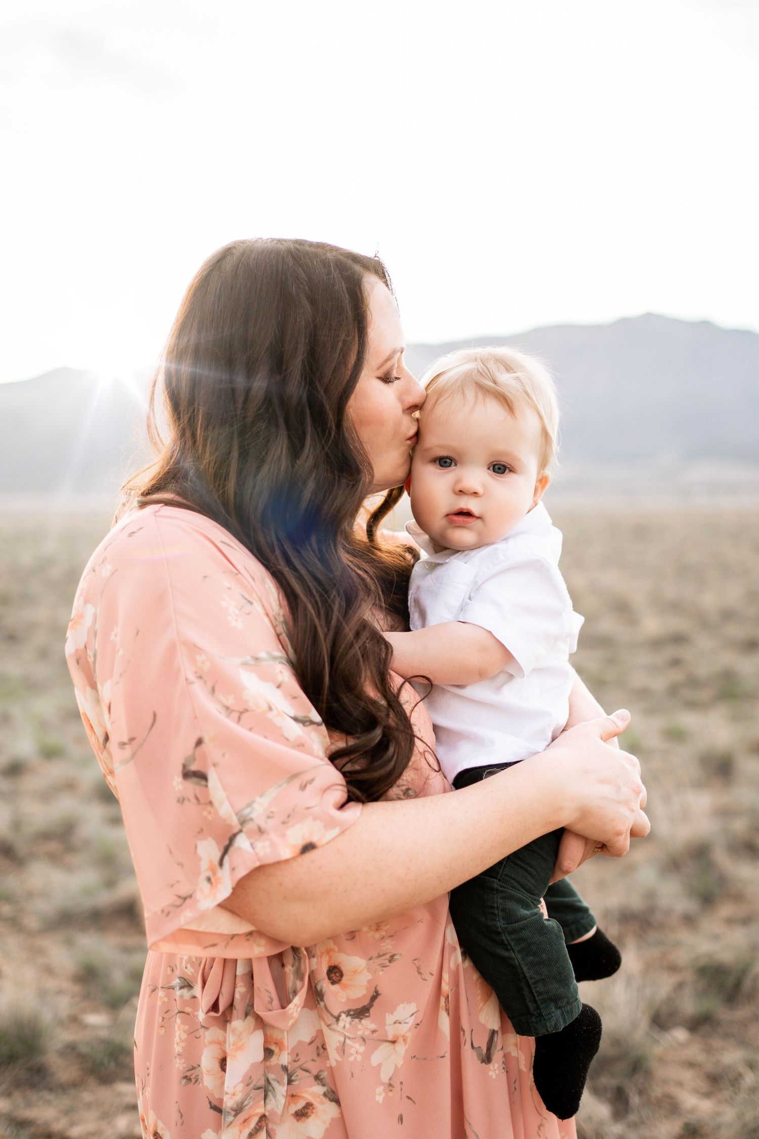 A mother in a floral pink dress kisses her baby boy outdoors with mountains in the background.