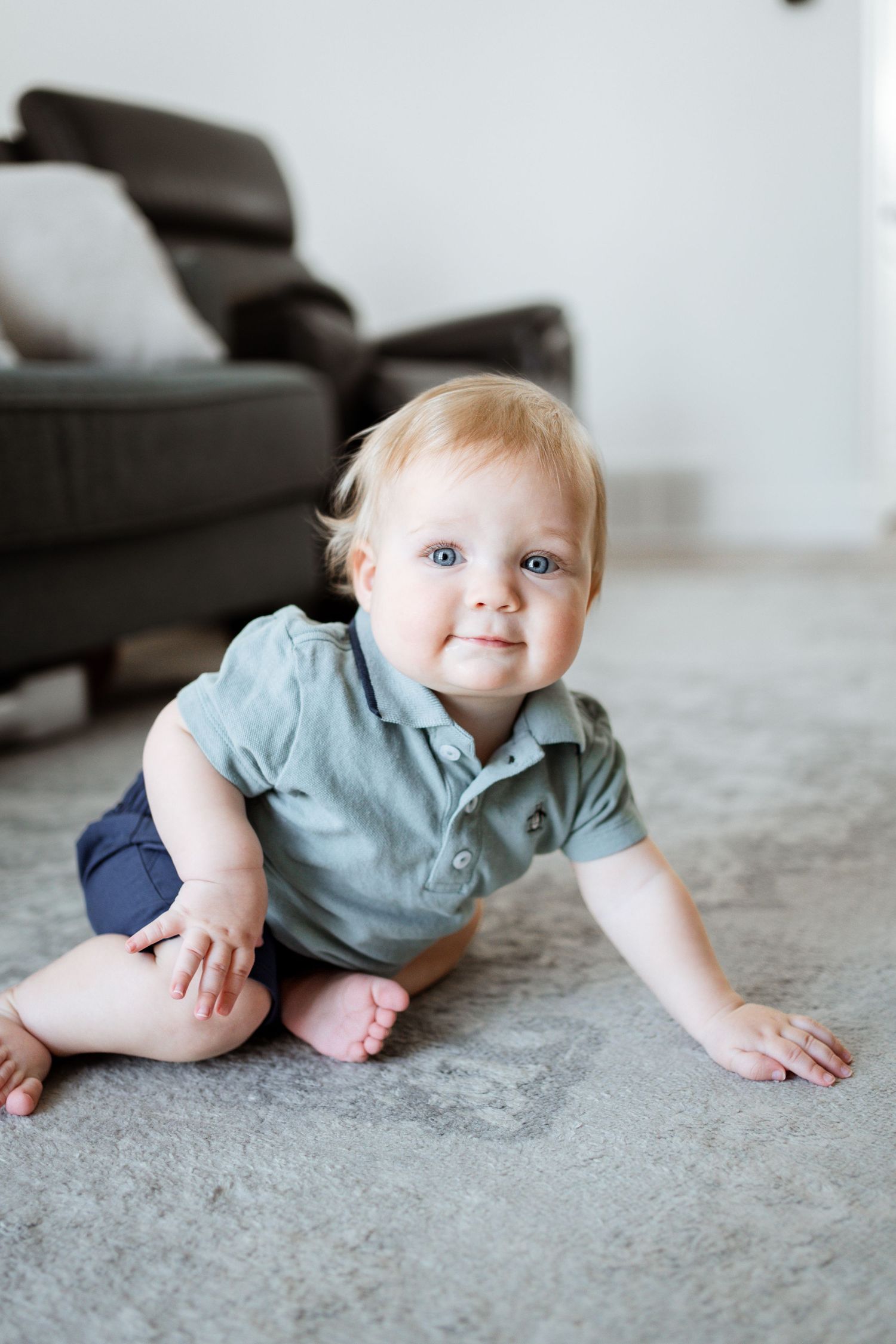 Baby in light blue polo shirt and navy shorts sits on gray carpet near dark couch.