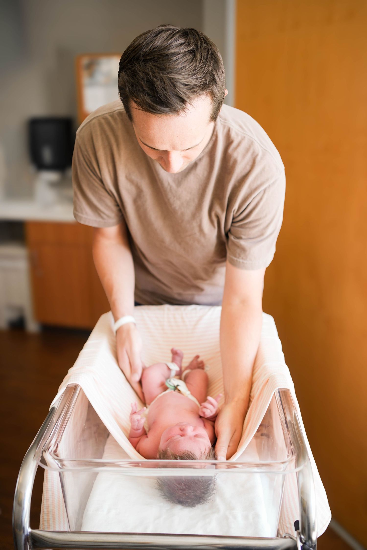 Father in hospital gown tenderly caring for newborn baby in bassinet after birth.