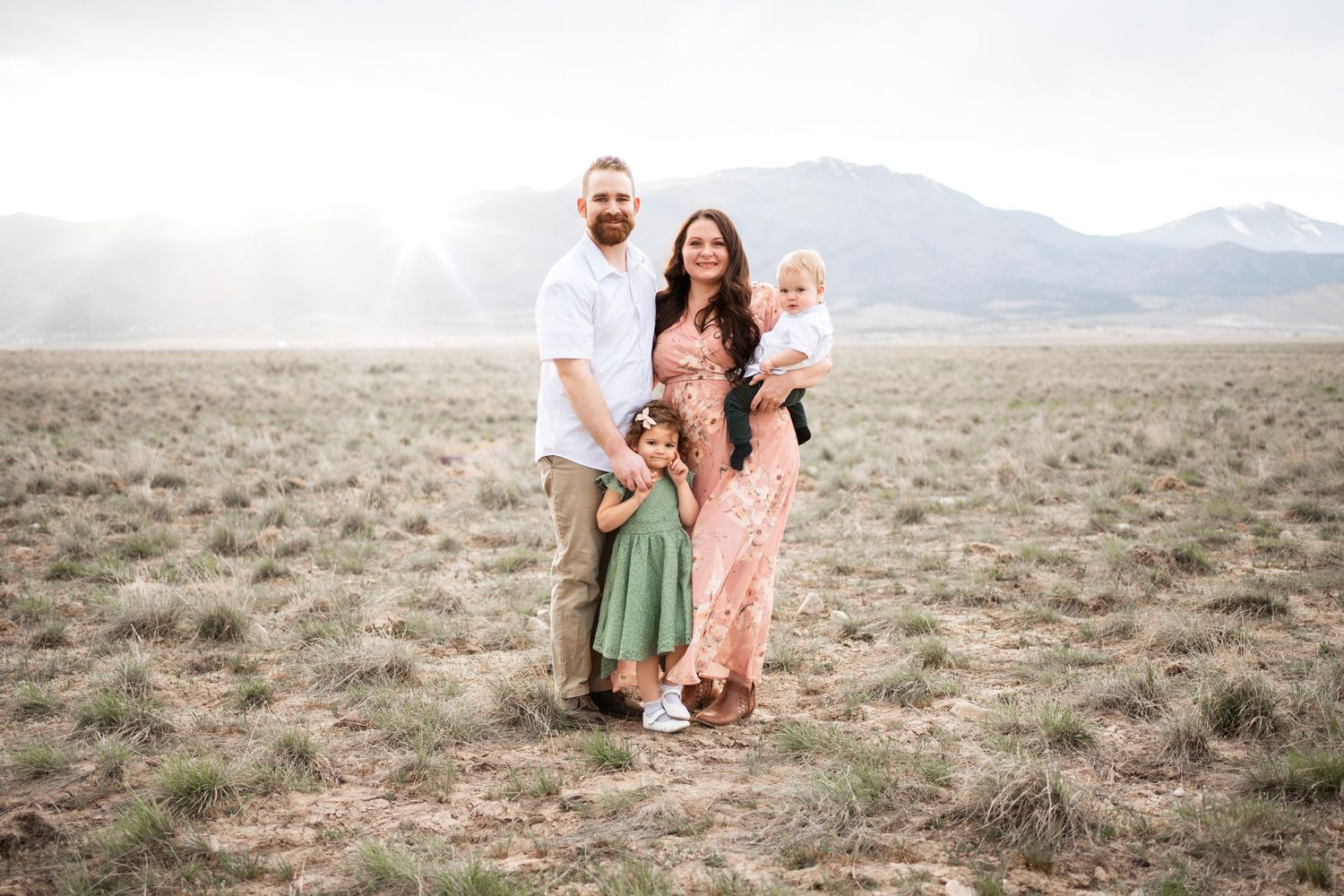 Family of four posing in a scenic mountain meadow, parents and two young children smiling outdoors.