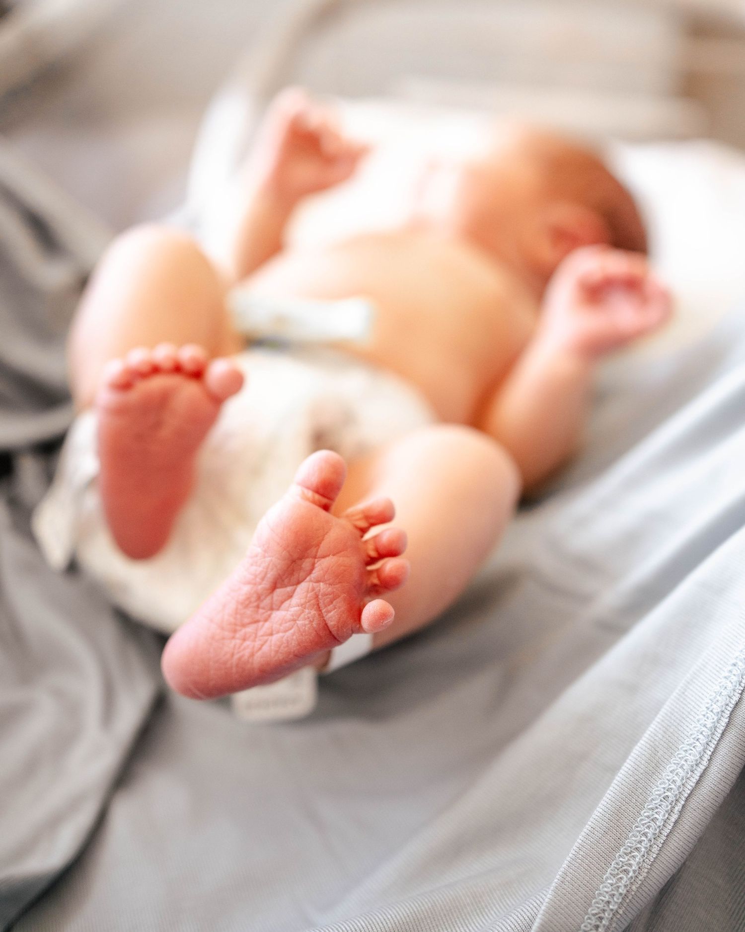 Close-up of a newborn baby's tiny feet and toes lying on a soft gray blanket in a diaper.