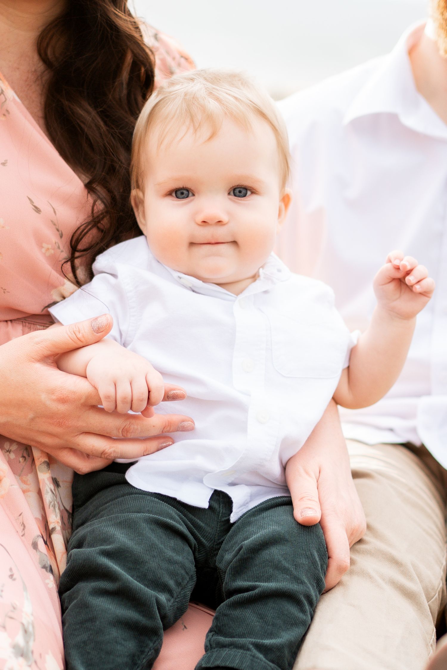Adorable blue-eyed baby in white shirt sitting on parents laps during a family portrait session.