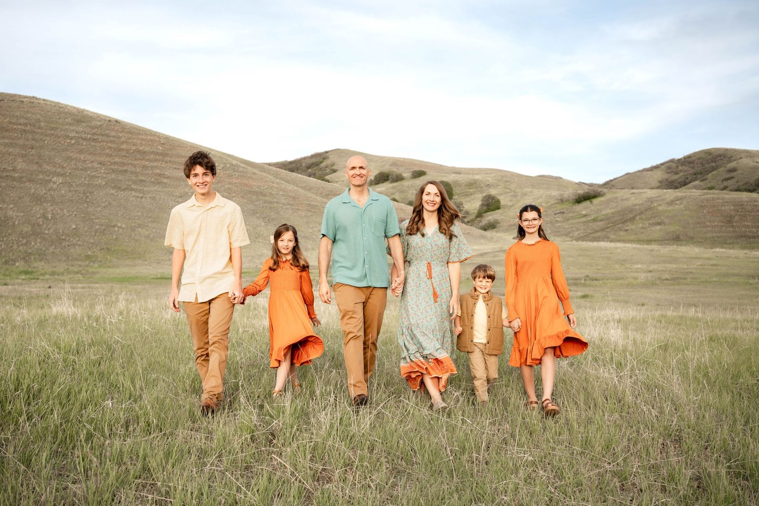 Family of six posing in a green meadow with rolling hills, wearing coordinated orange and teal outfits.