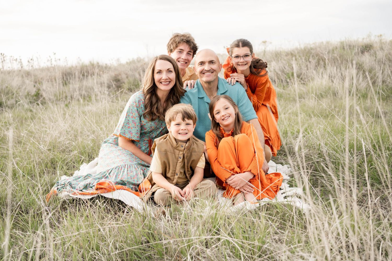 Smiling family of six in coordinated blue, orange and tan outfits posing together in a tall grassy meadow for an outdoor portrait.