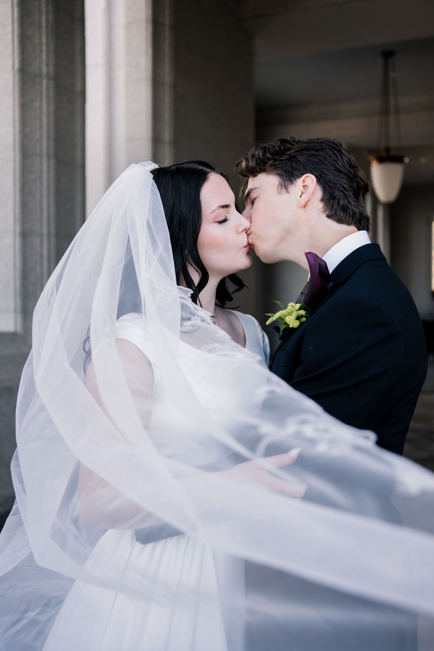 Bride in a flowing white veil kisses her groom in a black suit with a purple bow tie and green boutonniere beside stone columns.