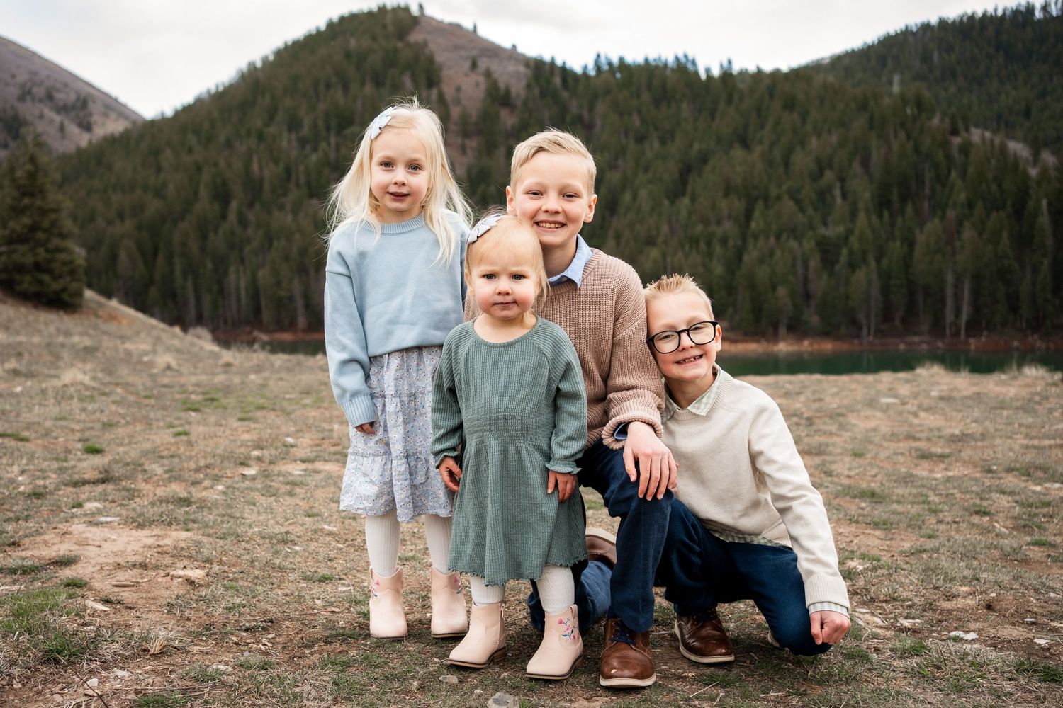 Family sits together on rocky ground with evergreen forest and mountains in background during outdoor photo session.