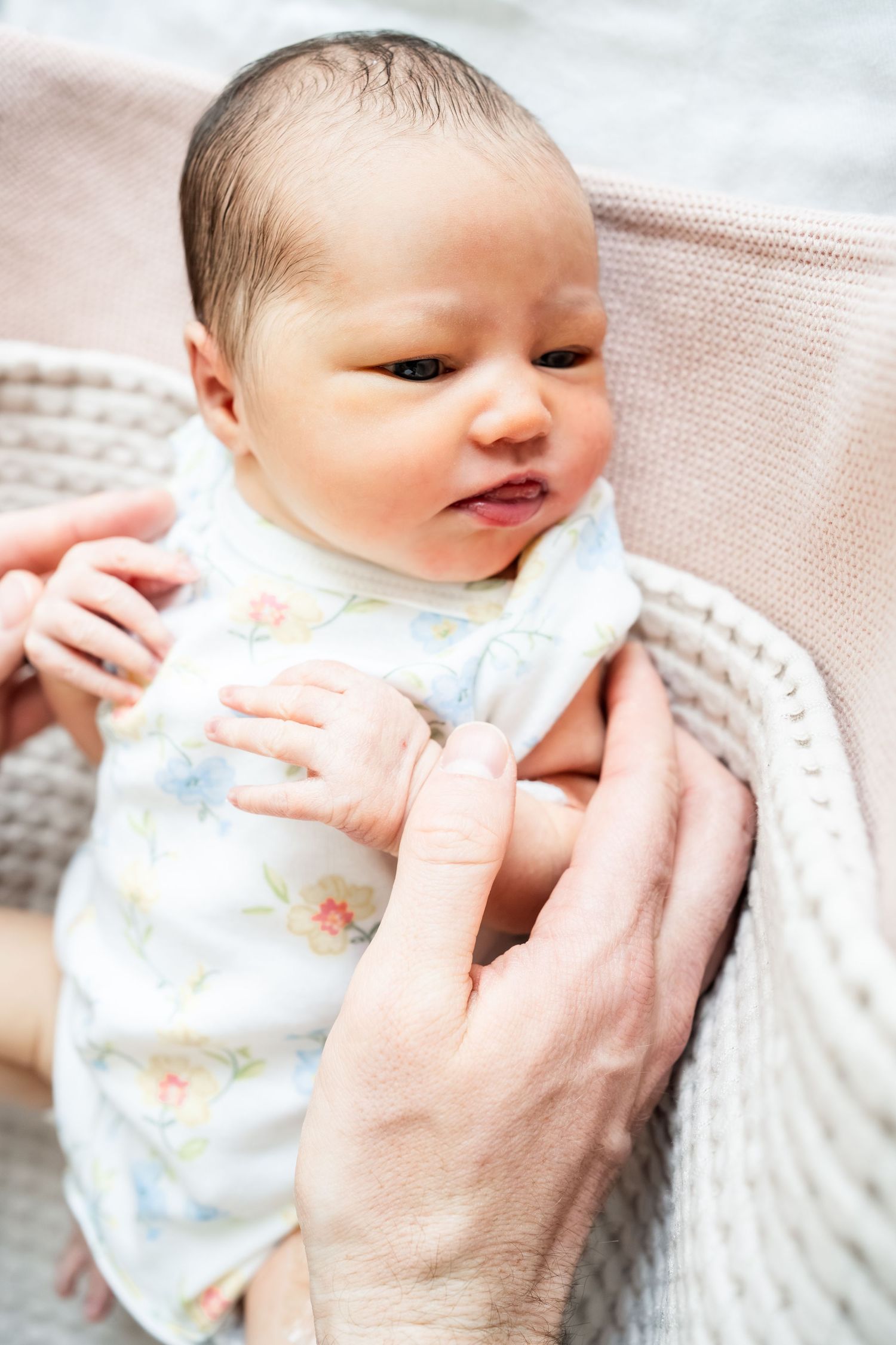 Close-up of a newborn baby in floral onesie being cradled in parent's arms with soft blanket.