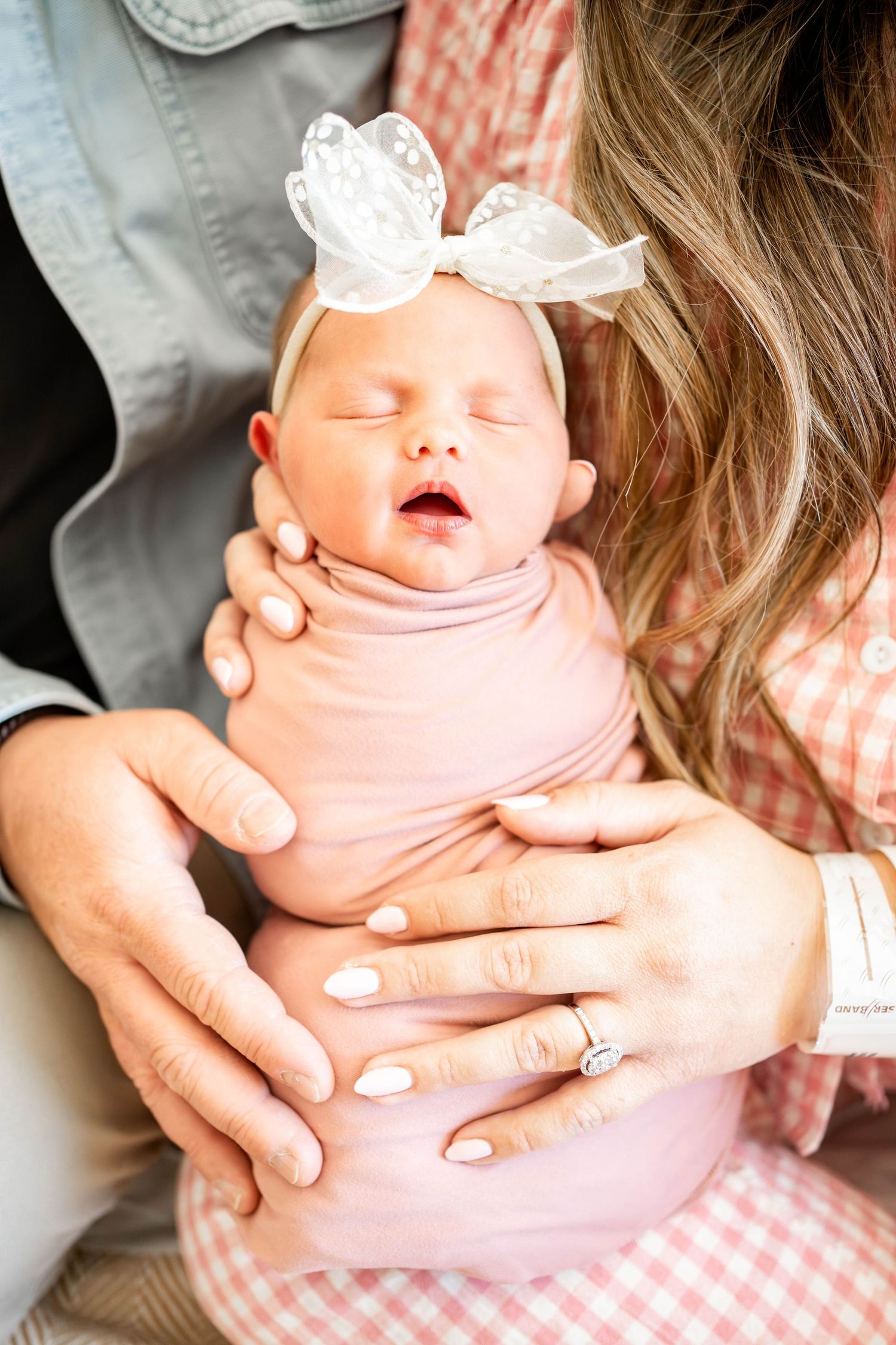 Newborn baby swaddled in pink with a white bow headband, cradled in parents' arms during a newborn photo session.