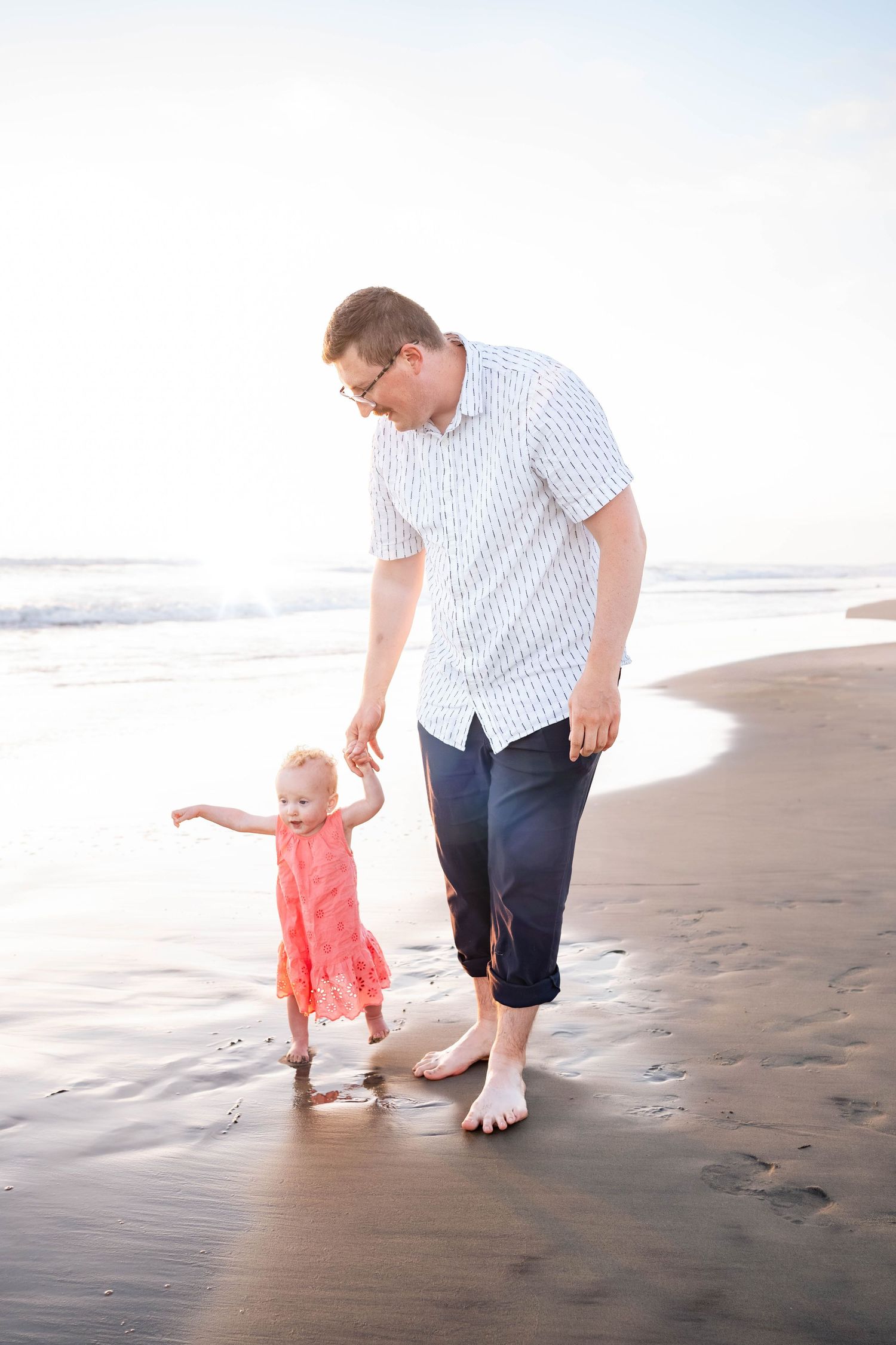 Father helps toddler girl in pink dress take first steps on sandy beach with ocean waves in background.
