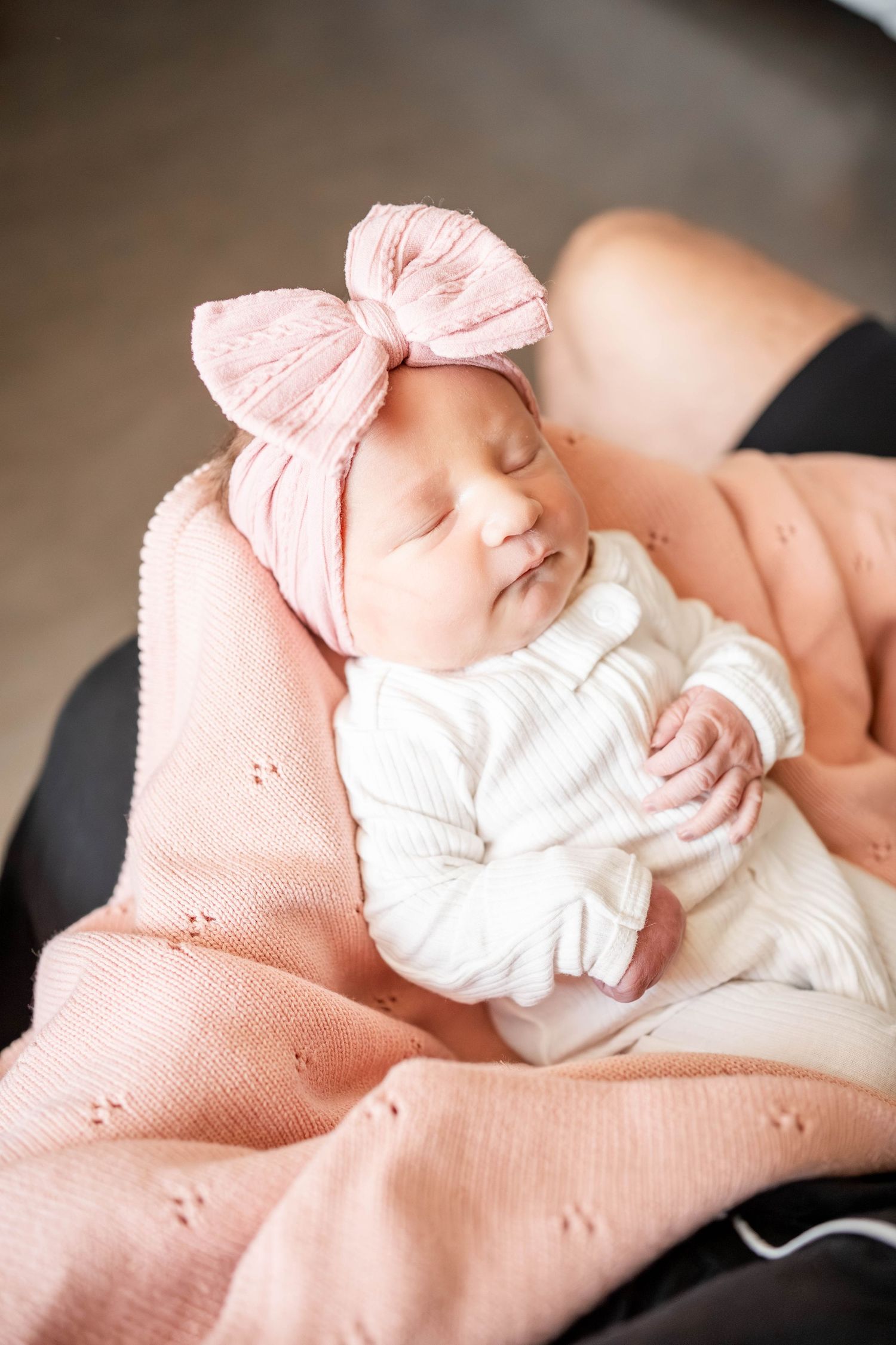 Sleeping newborn baby in white striped onesie and pink bow headband, resting on a soft pink blanket.