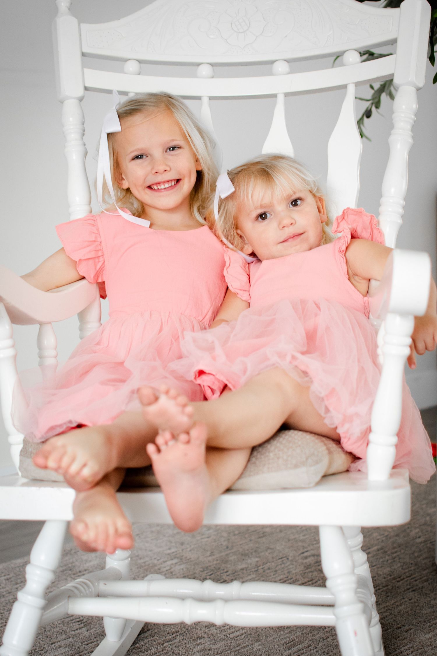 Two children in pink tulle dresses sit playfully on a white wooden chair.