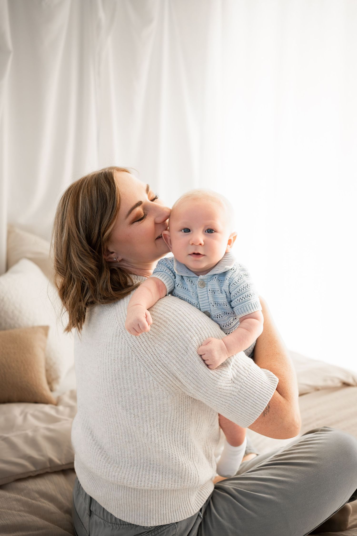 Smiling mother in a cream knit sweater kisses her baby boy in a light blue shirt while sitting on a bed with soft white curtains.