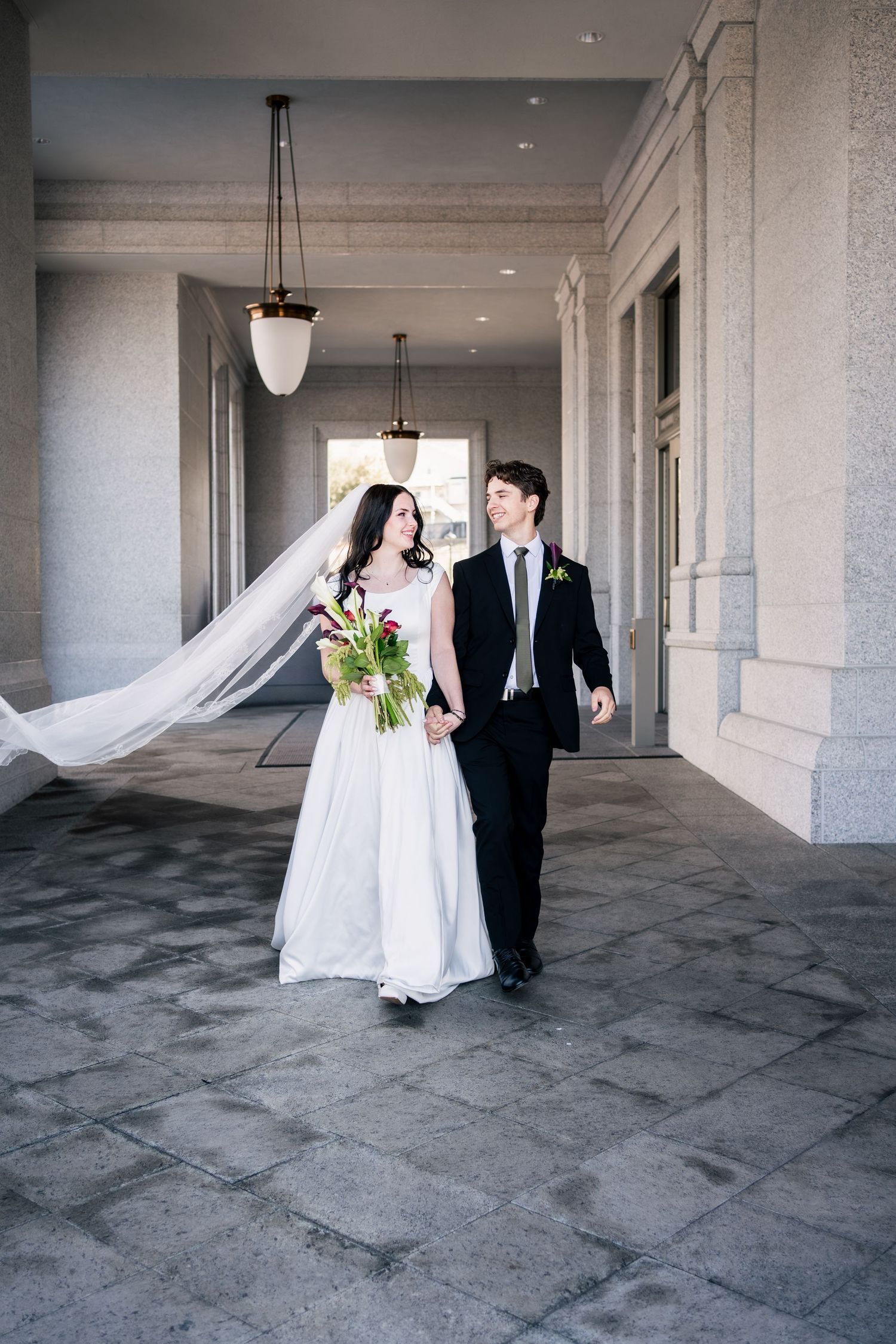 Bride in a white gown with a long flowing veil holds hands with a groom in a black suit while walking under a stone covered walkway.