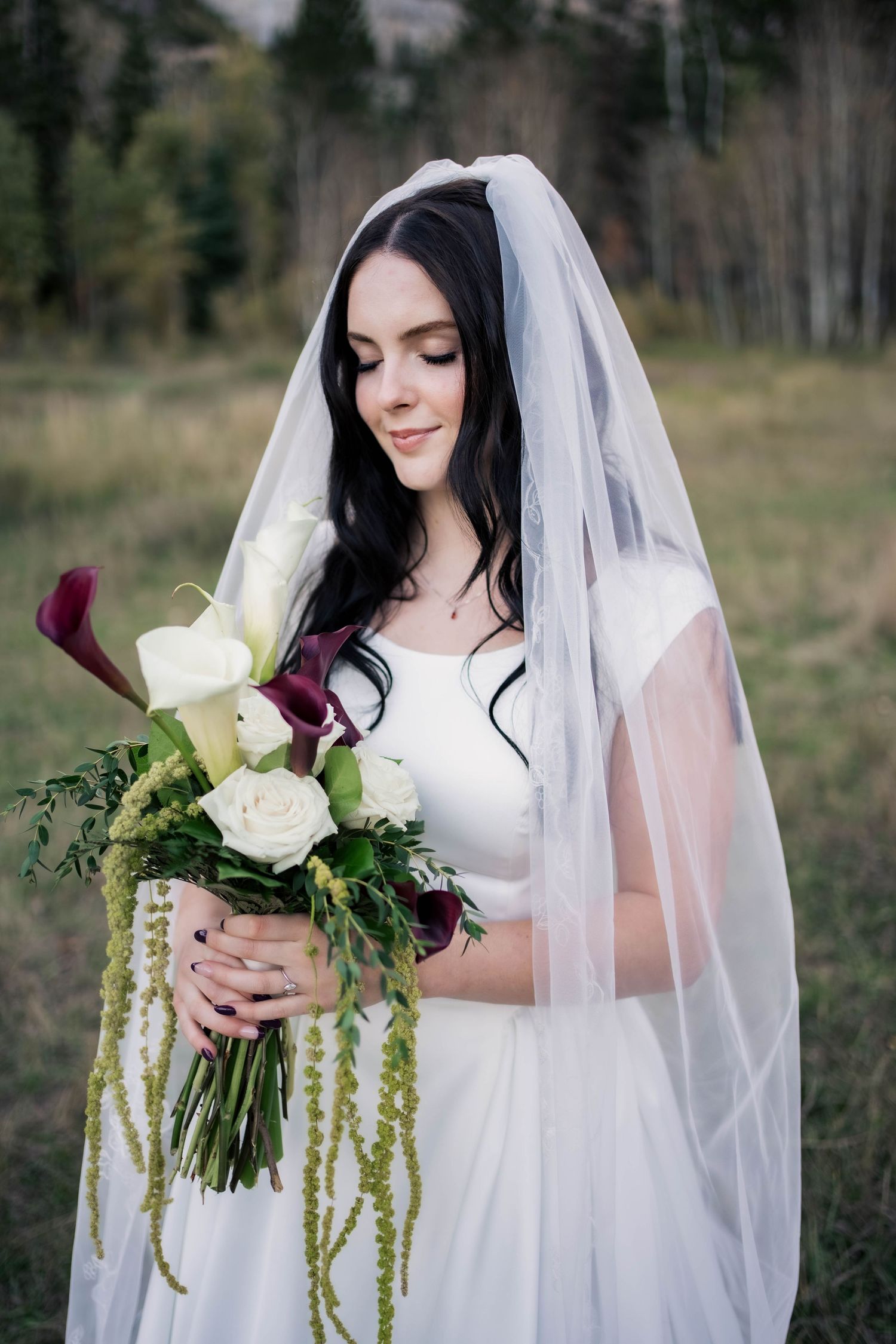 Bride in a white gown and long sheer veil looks down at her bouquet of white and burgundy calla lilies with trailing greenery in a meadow.