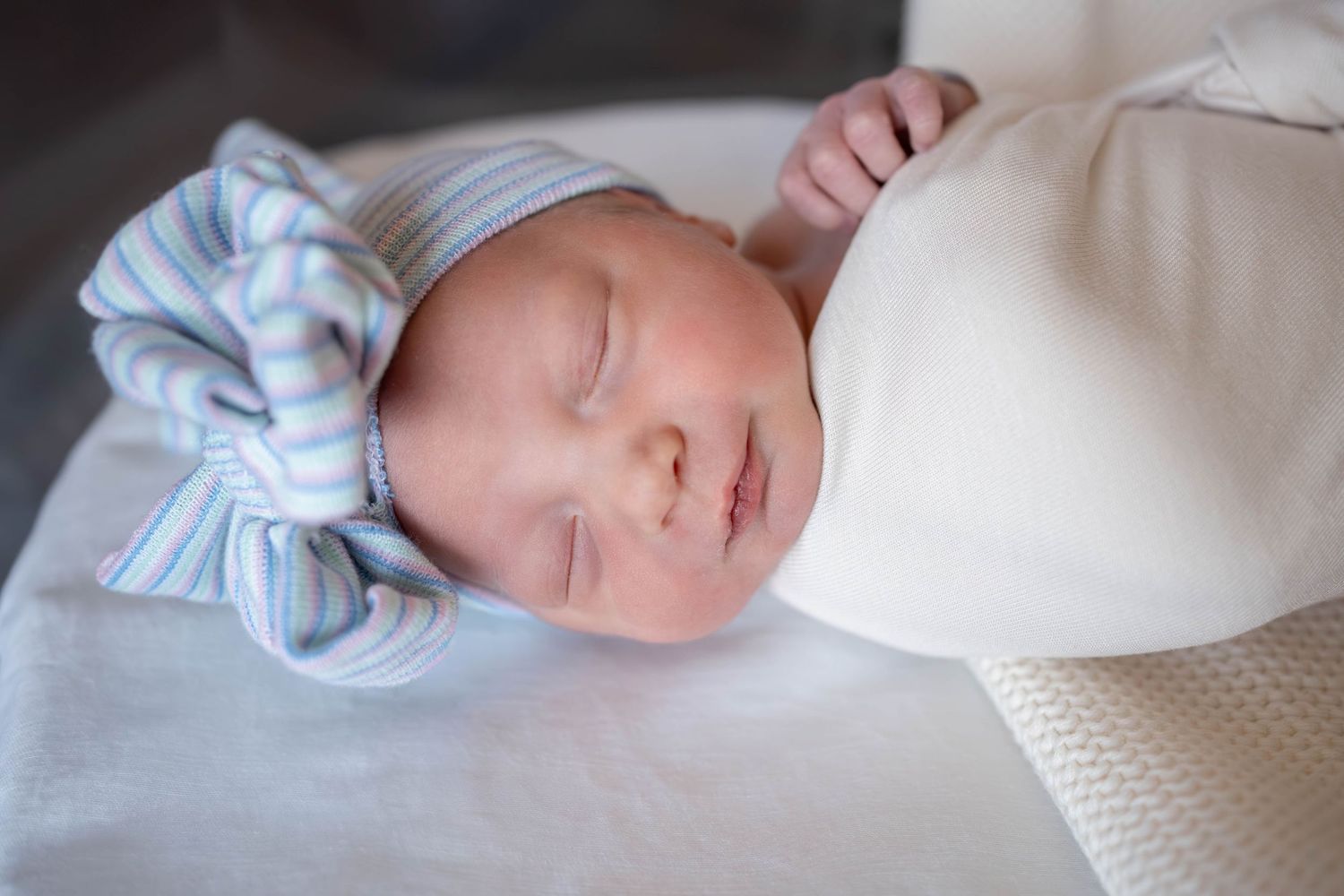 Sleeping newborn baby wrapped in a cream swaddle blanket wearing a blue and white striped knotted hospital-style hat.