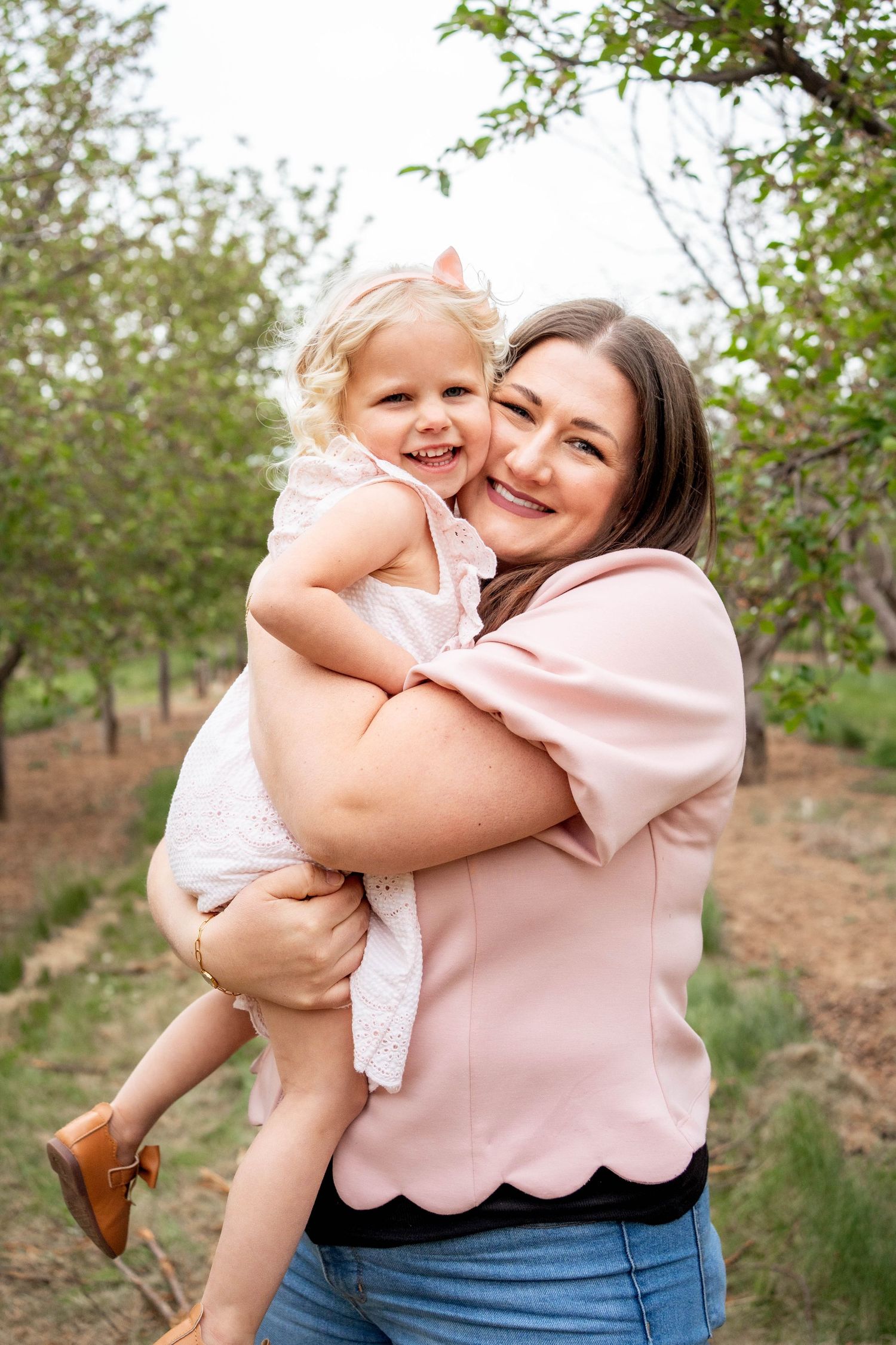 A smiling mother in a pink top holds her laughing toddler daughter in a white dress outdoors among green trees.