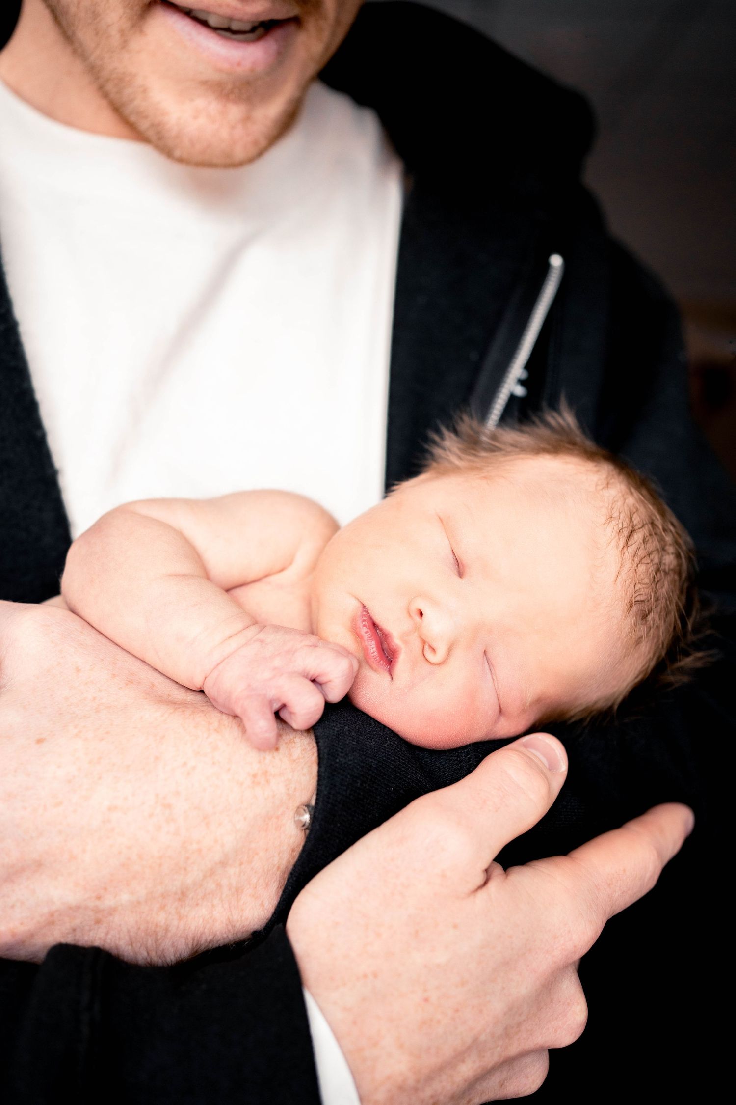 Father tenderly holding sleeping newborn baby wrapped in black blanket shortly after birth.