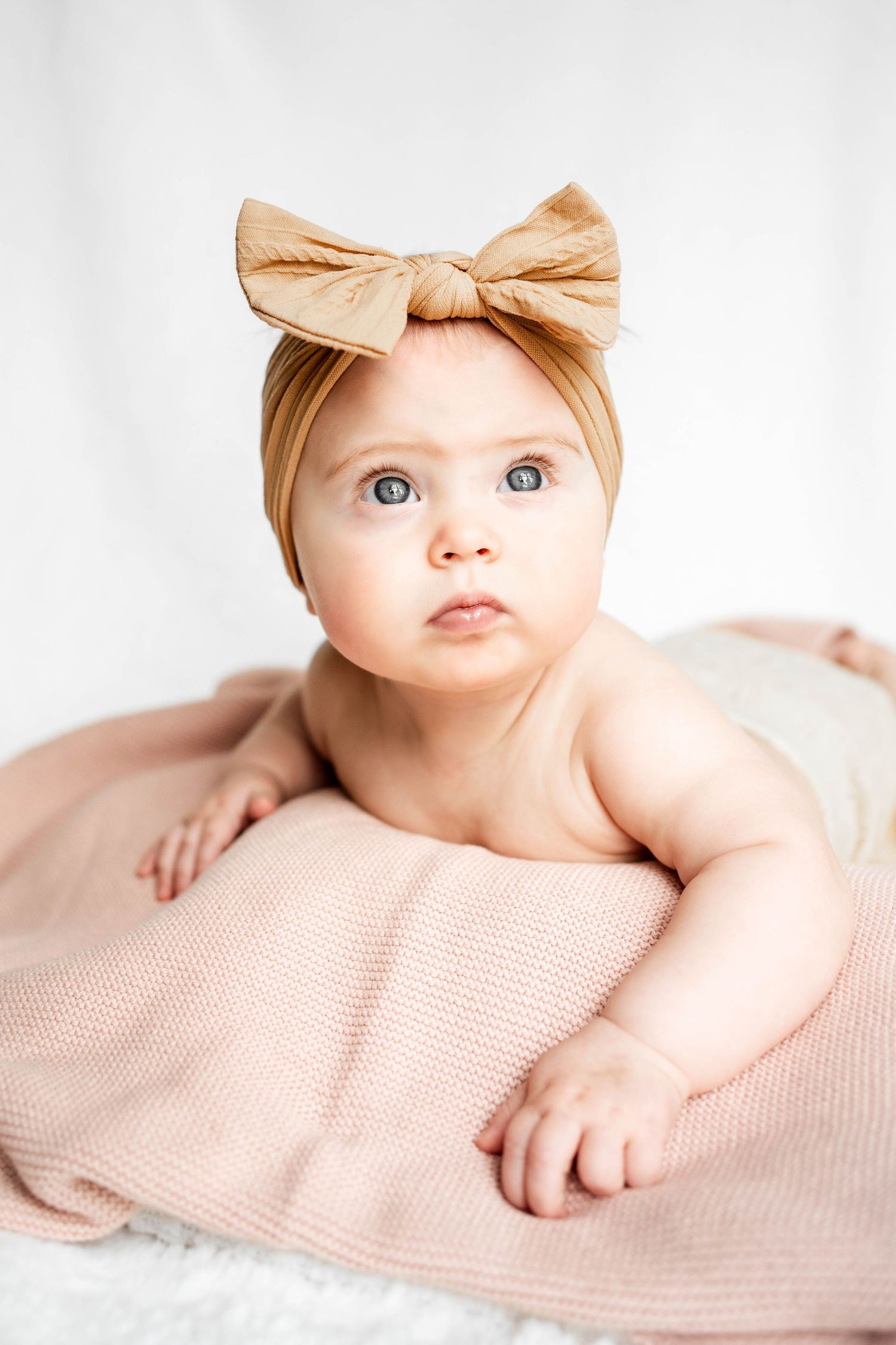 Adorable baby girl wearing a tan bow headband lying on a pink knit blanket against a white background.