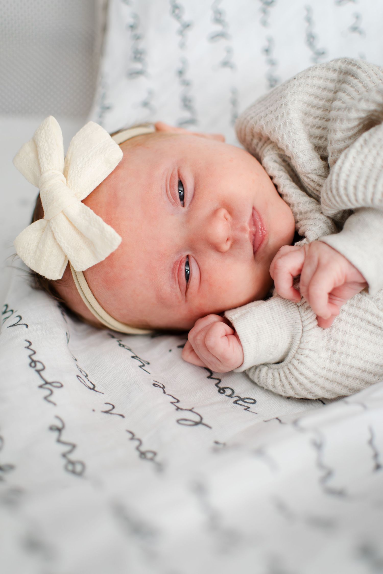 A tiny baby wearing a cream knit sweater and white bow headband rests on patterned fabric.