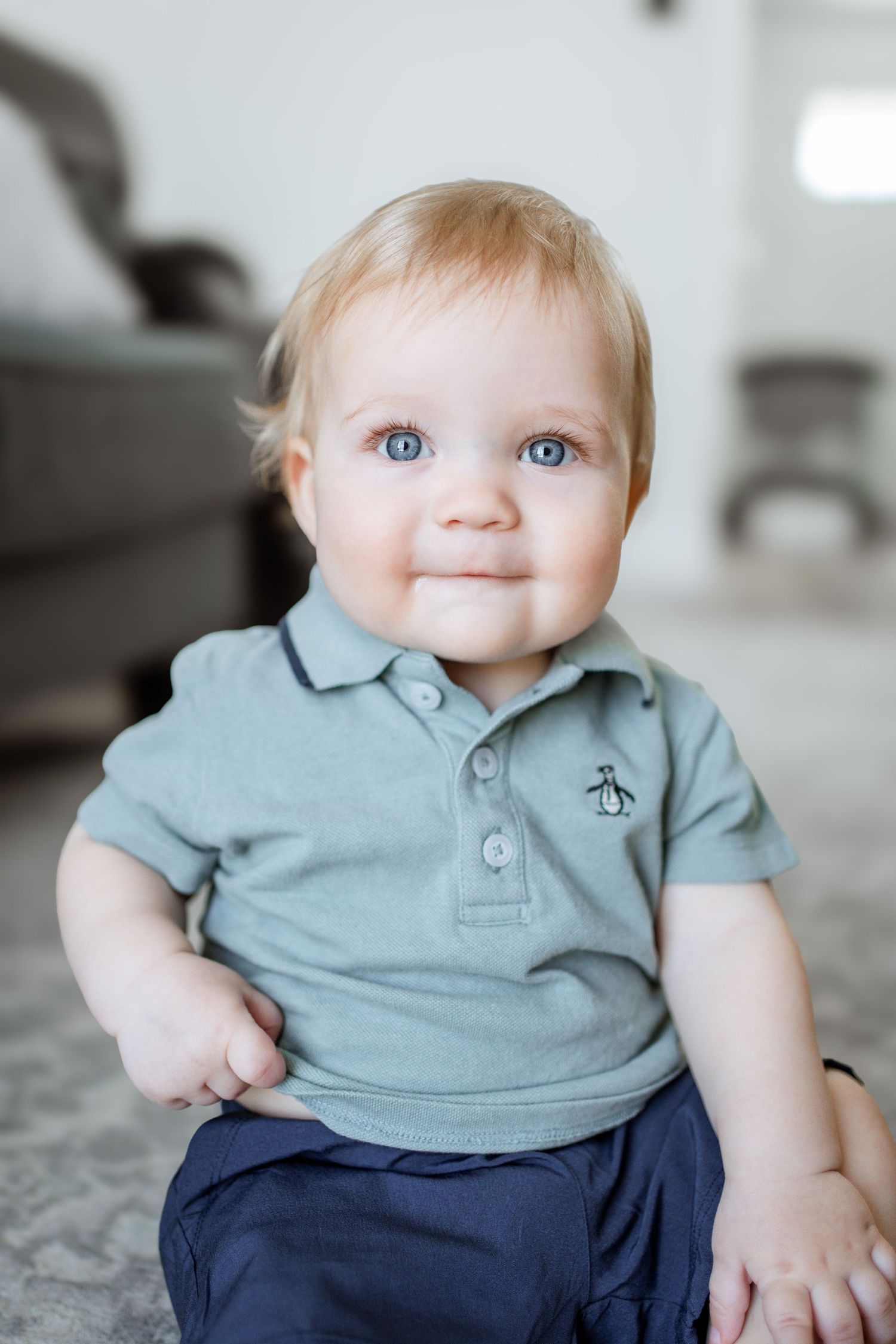 A baby wearing a light blue polo shirt with a small emblem and navy pants sits on a gray carpet.