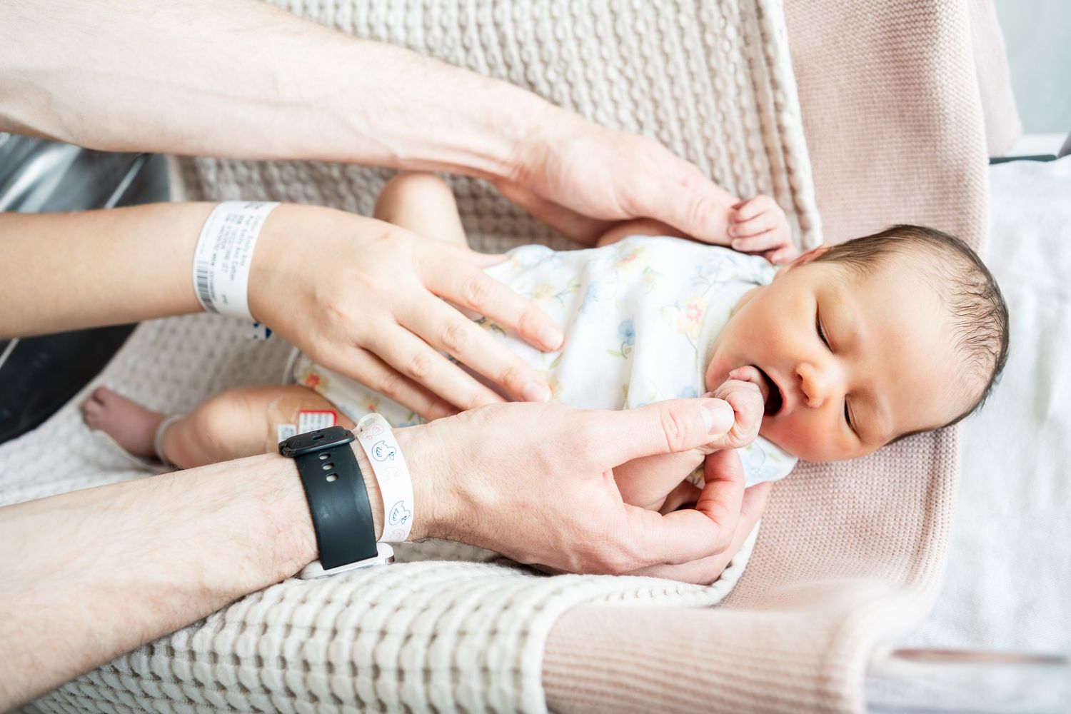 Newborn baby sleeping peacefully while parents gently hold hands together in a hospital setting.