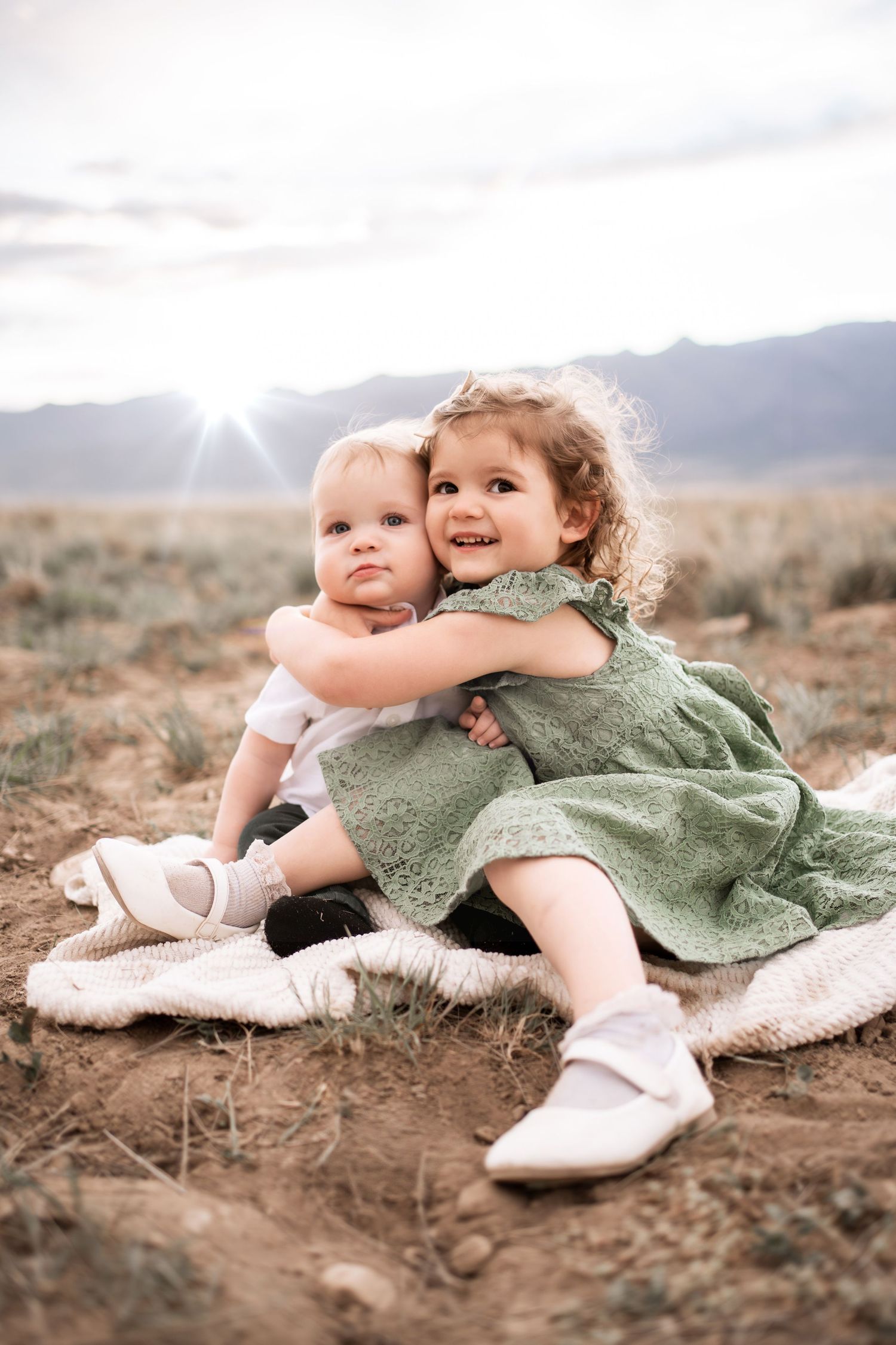 Two young sisters sitting on a blanket in a desert field, older girl in green dress hugging baby sibling.