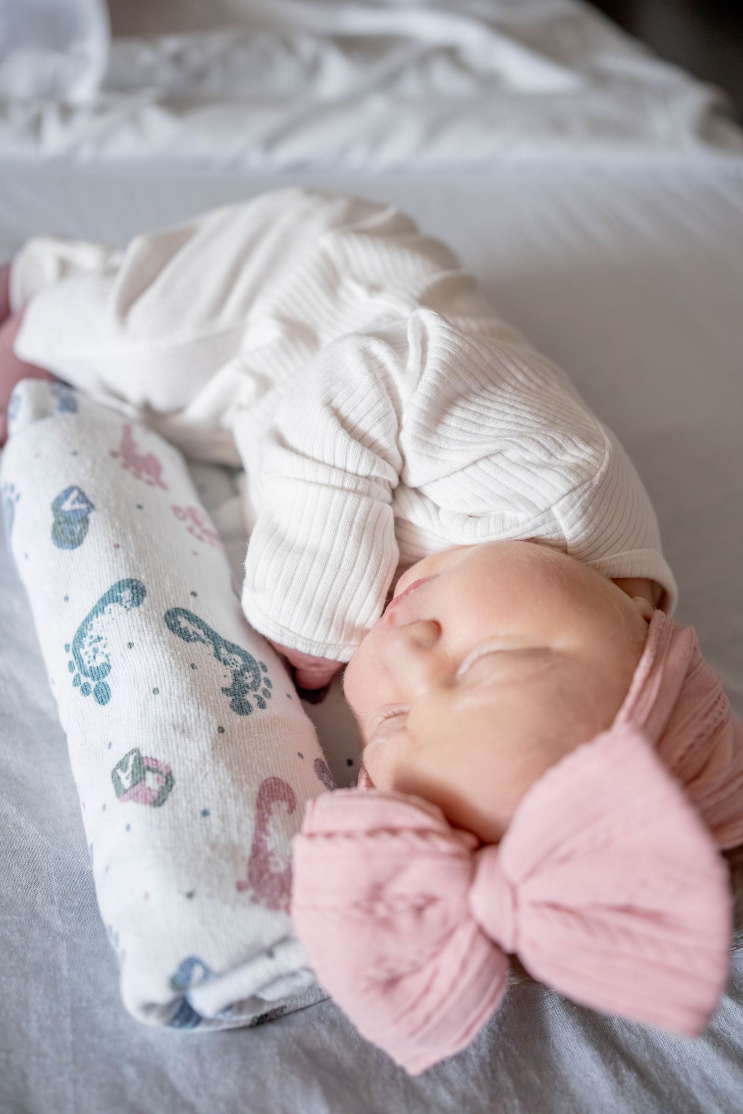 Sleeping newborn baby girl wearing a pink bow headband and white ribbed outfit, wrapped in a floral swaddle blanket.