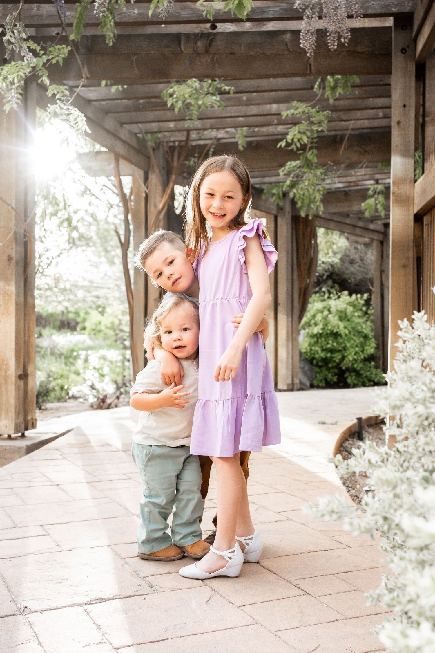 Three young children pose together under a wooden pergola in a garden, with the oldest girl wearing a purple dress.