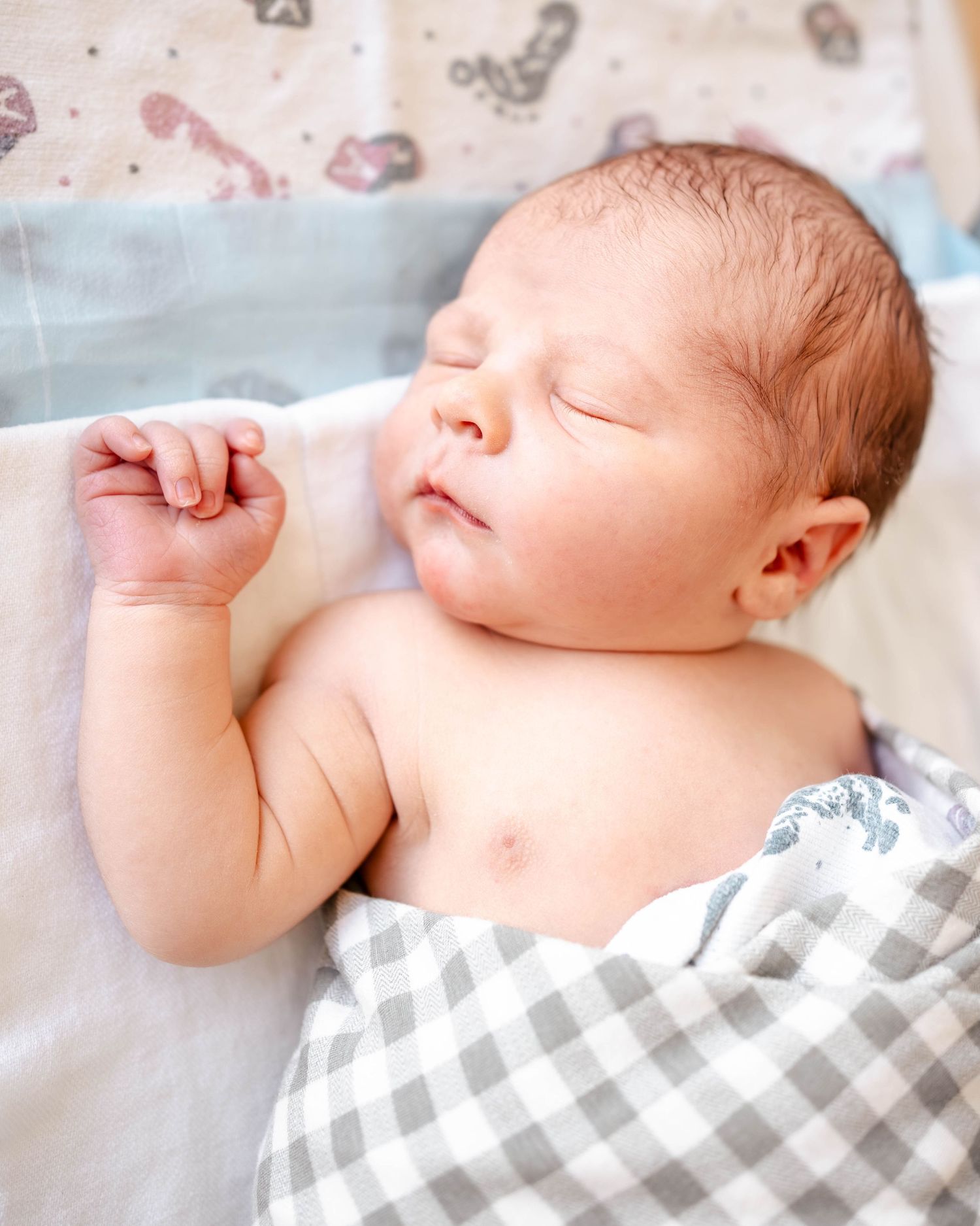 Peaceful newborn baby sleeping swaddled in gray gingham blanket, tiny fist raised, soft natural light photography.
