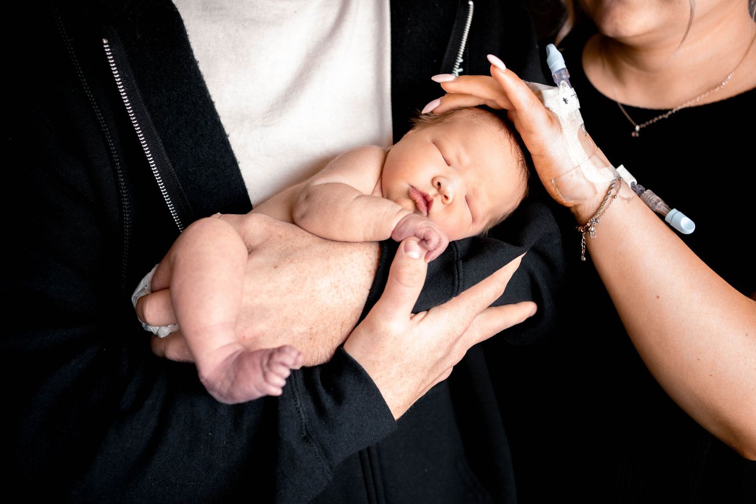 Newborn baby sleeping peacefully in parent's arms while another hand gently touches the infant's head.