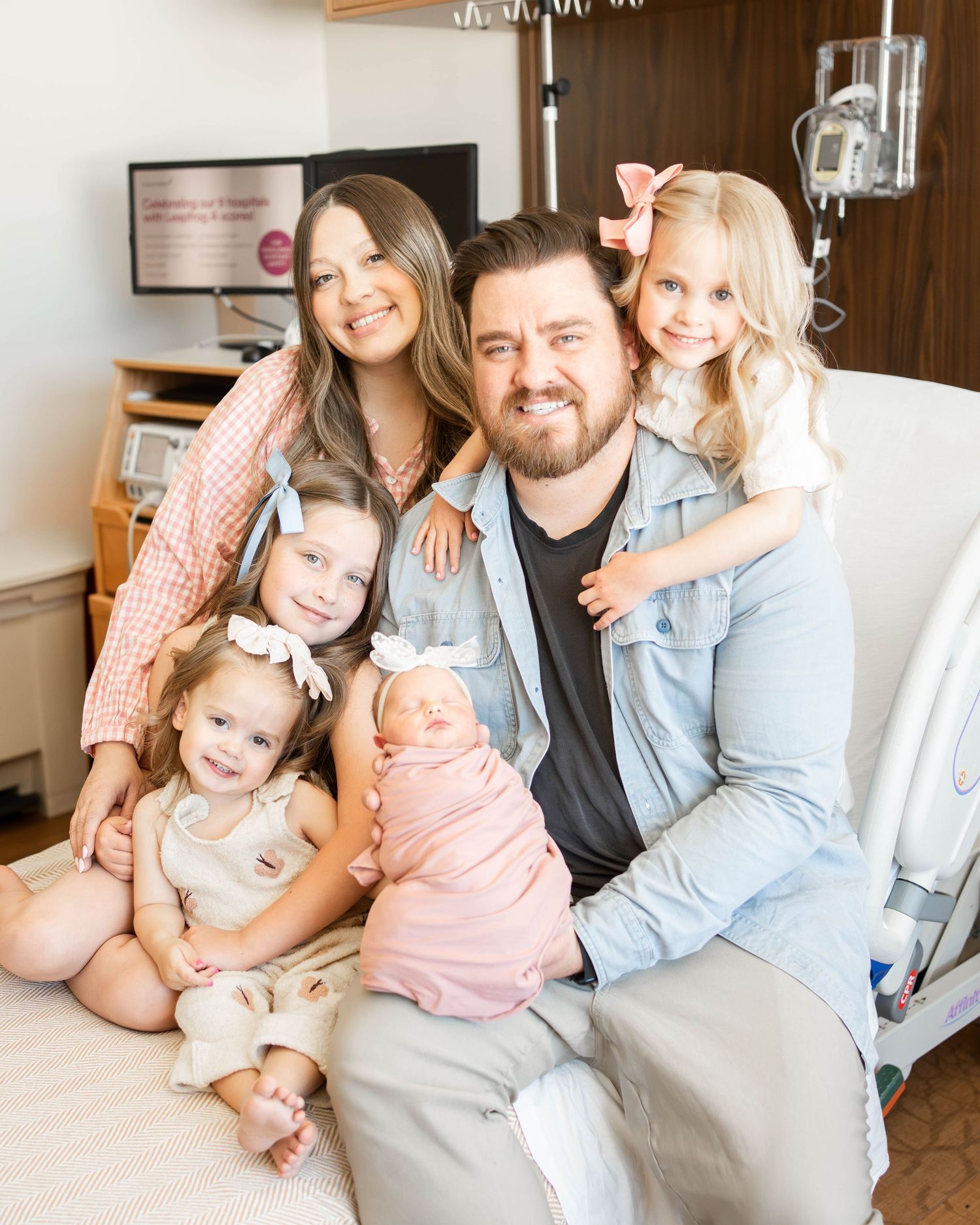 A family of six poses in a hospital room, parents with four young daughters including a newborn wrapped in pink.