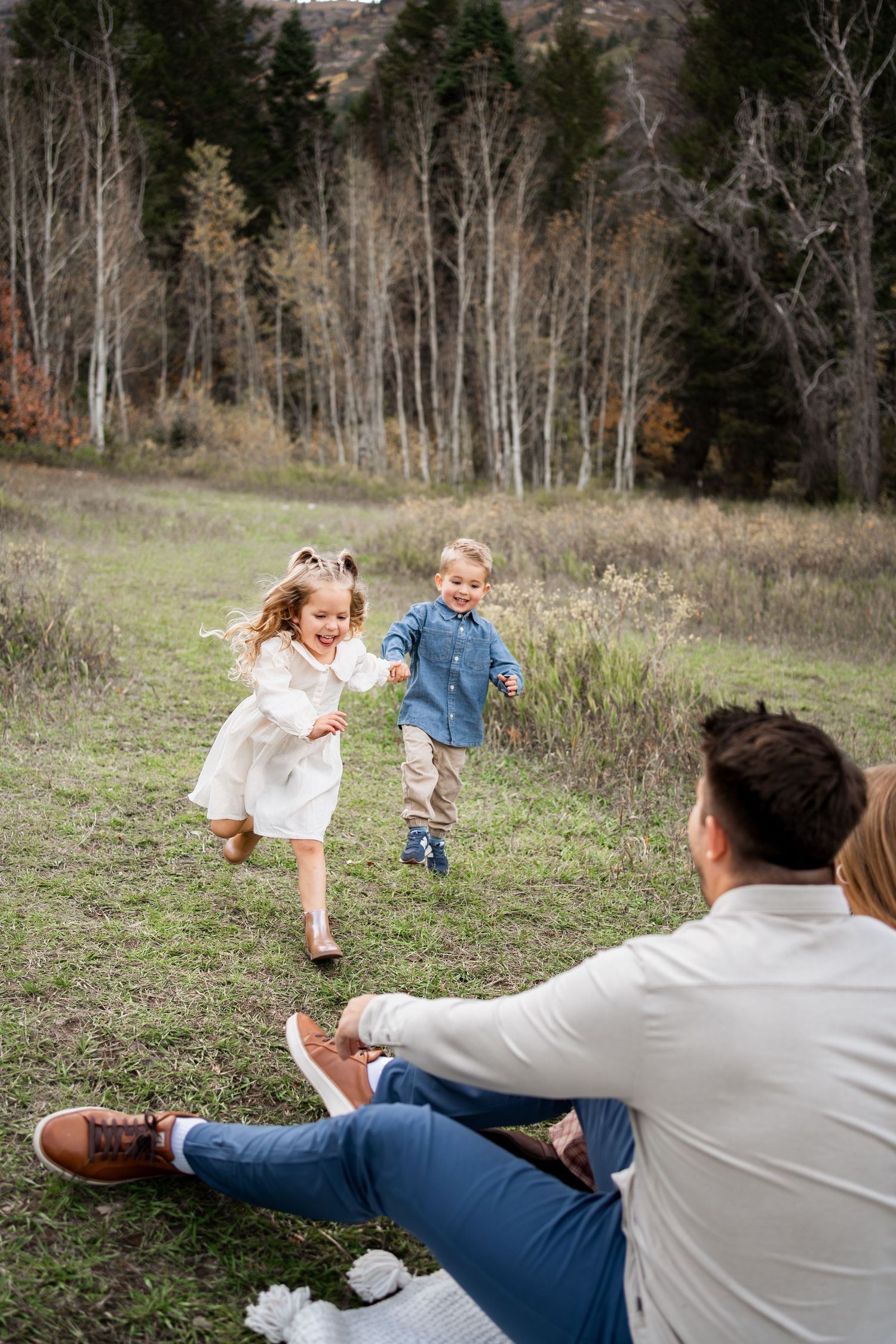 Two young children hold hands and run joyfully across a grassy meadow toward seated parents, with autumn aspen trees in the background.