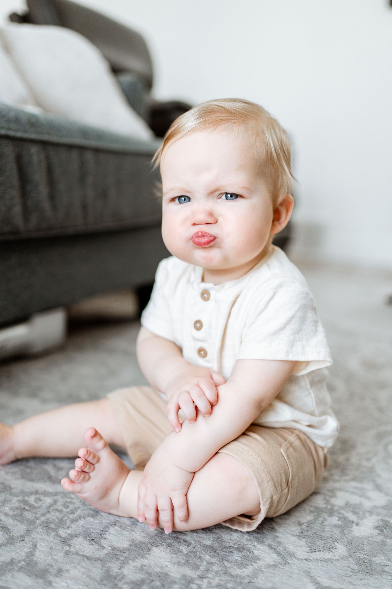 Cute baby with pouty lips sits on carpet in white shirt and tan shorts, making an adorable grumpy face.
