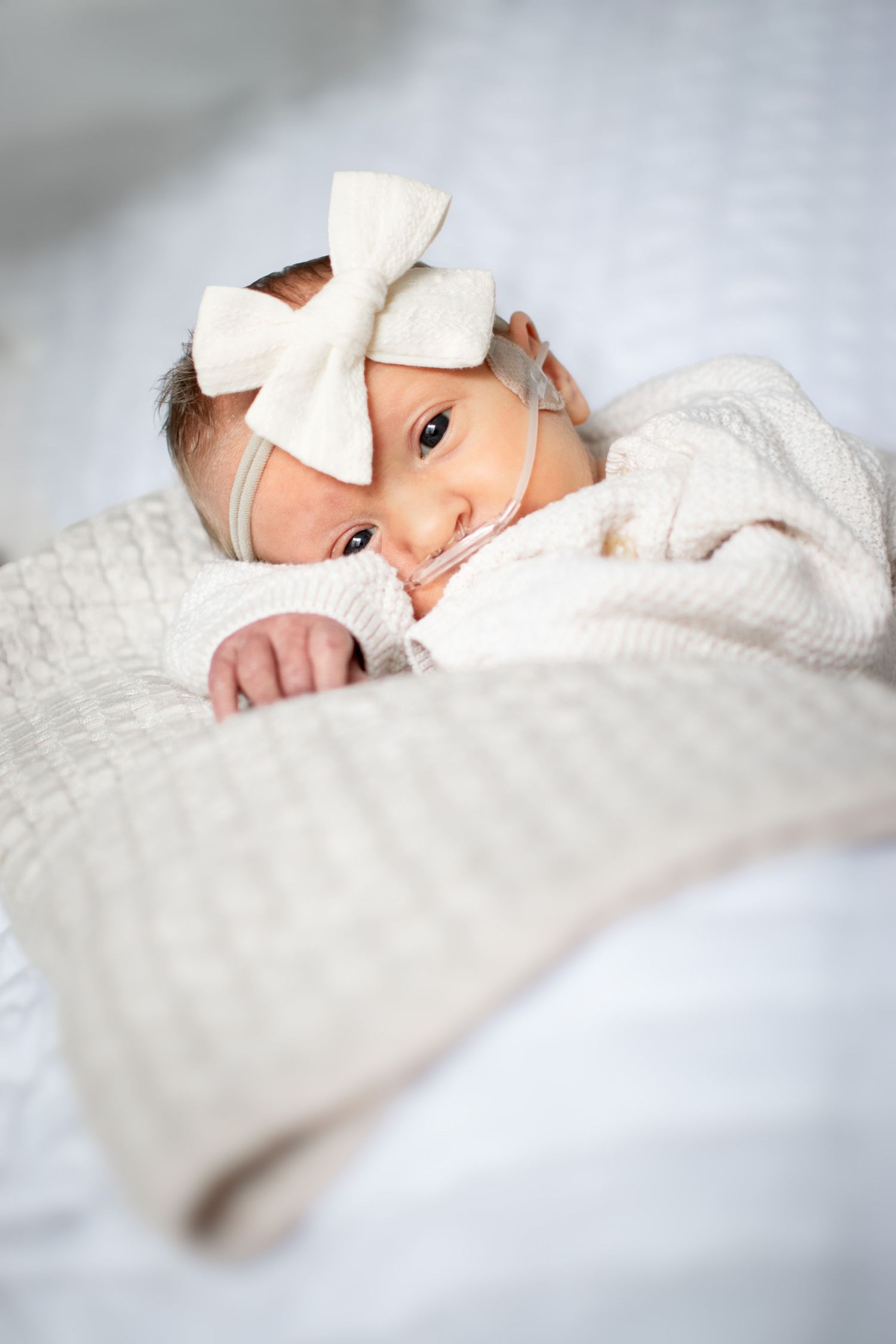 A baby wearing a white bow lies peacefully on soft white bedding in a series of dreamy, ethereal portraits.