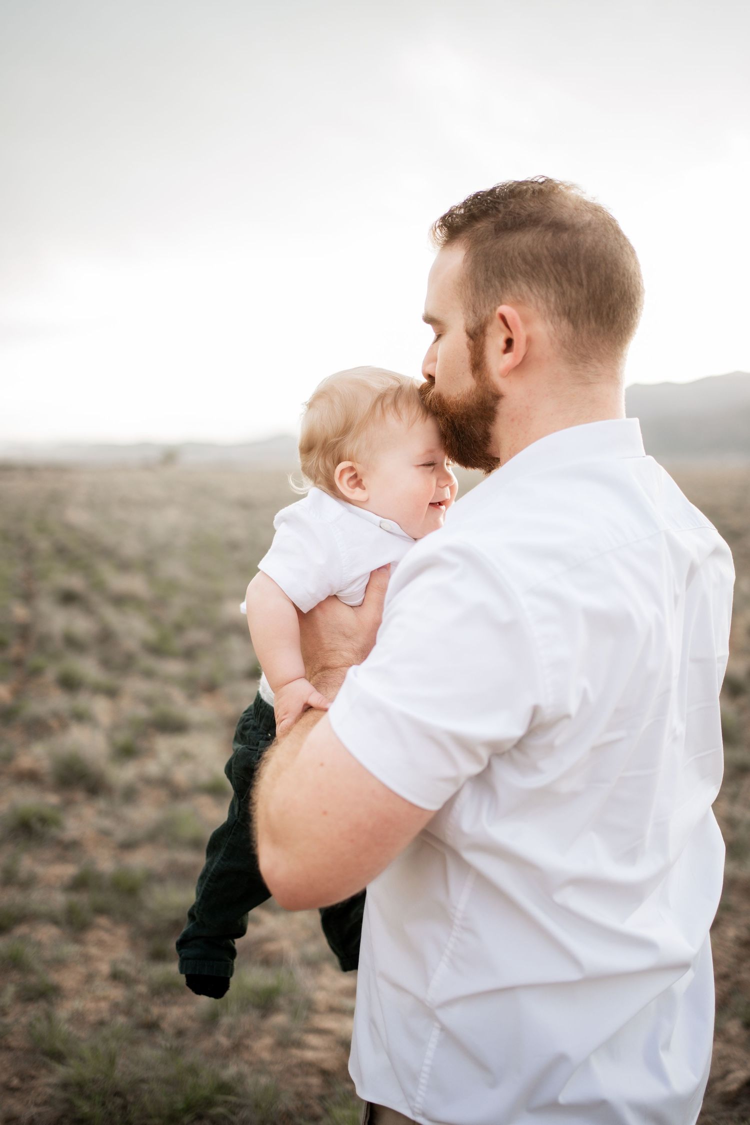 Bearded father in white shirt tenderly holds infant baby outdoors in open field landscape.