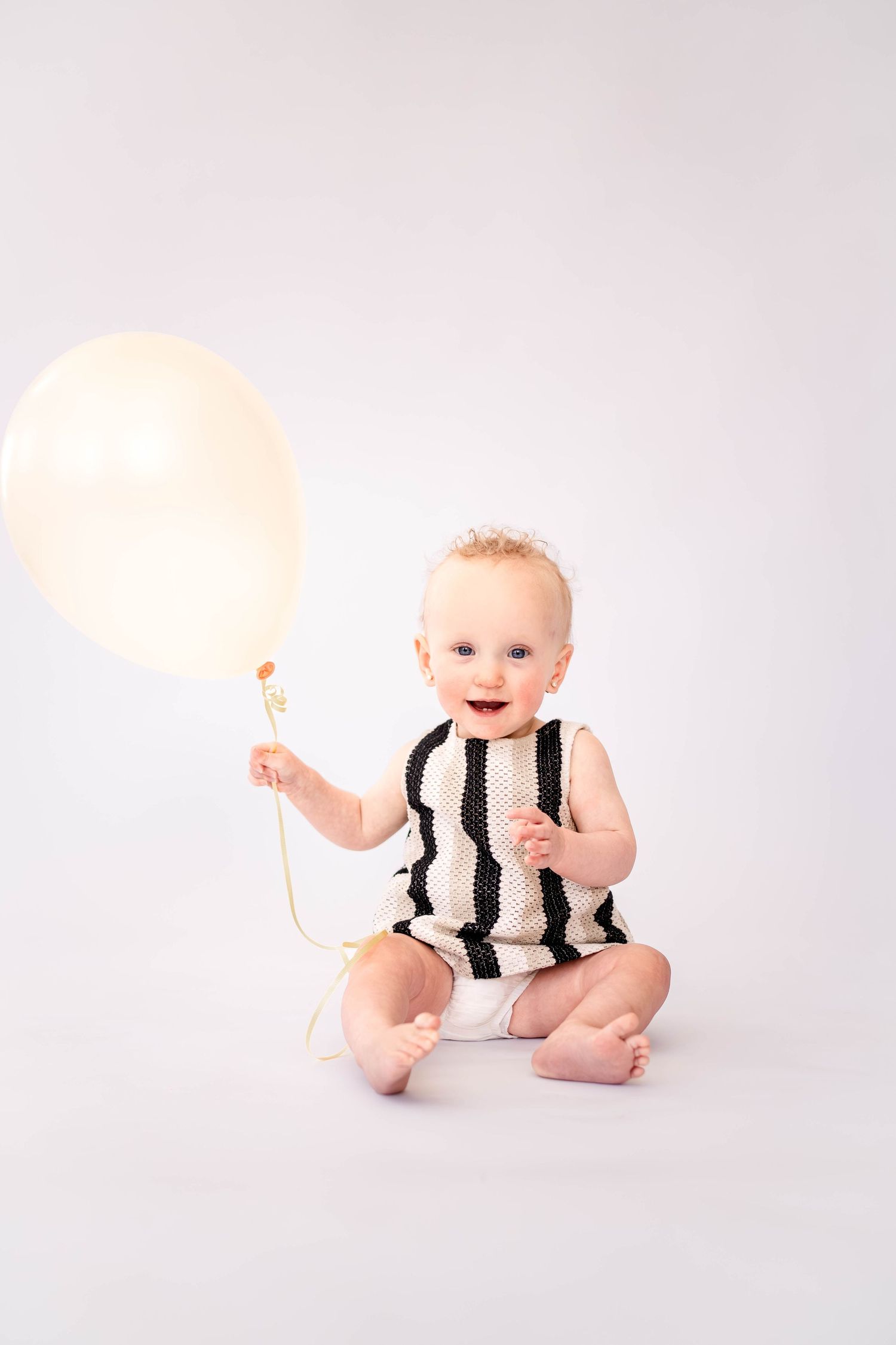Smiling baby in black and white striped outfit sitting on white background holding a cream balloon.