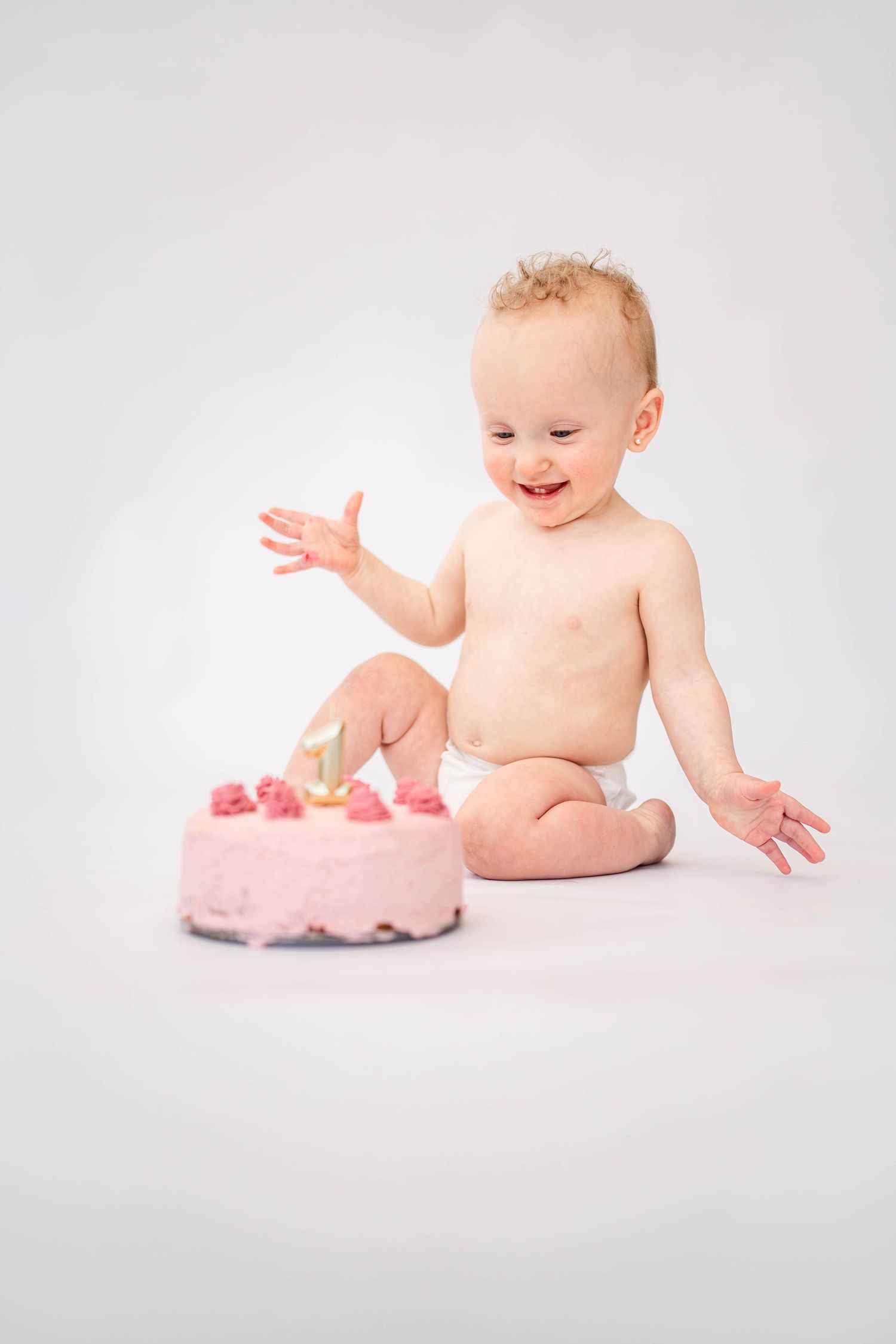 A happy baby in a diaper sits beside a pink first birthday cake with a candle on a white background.