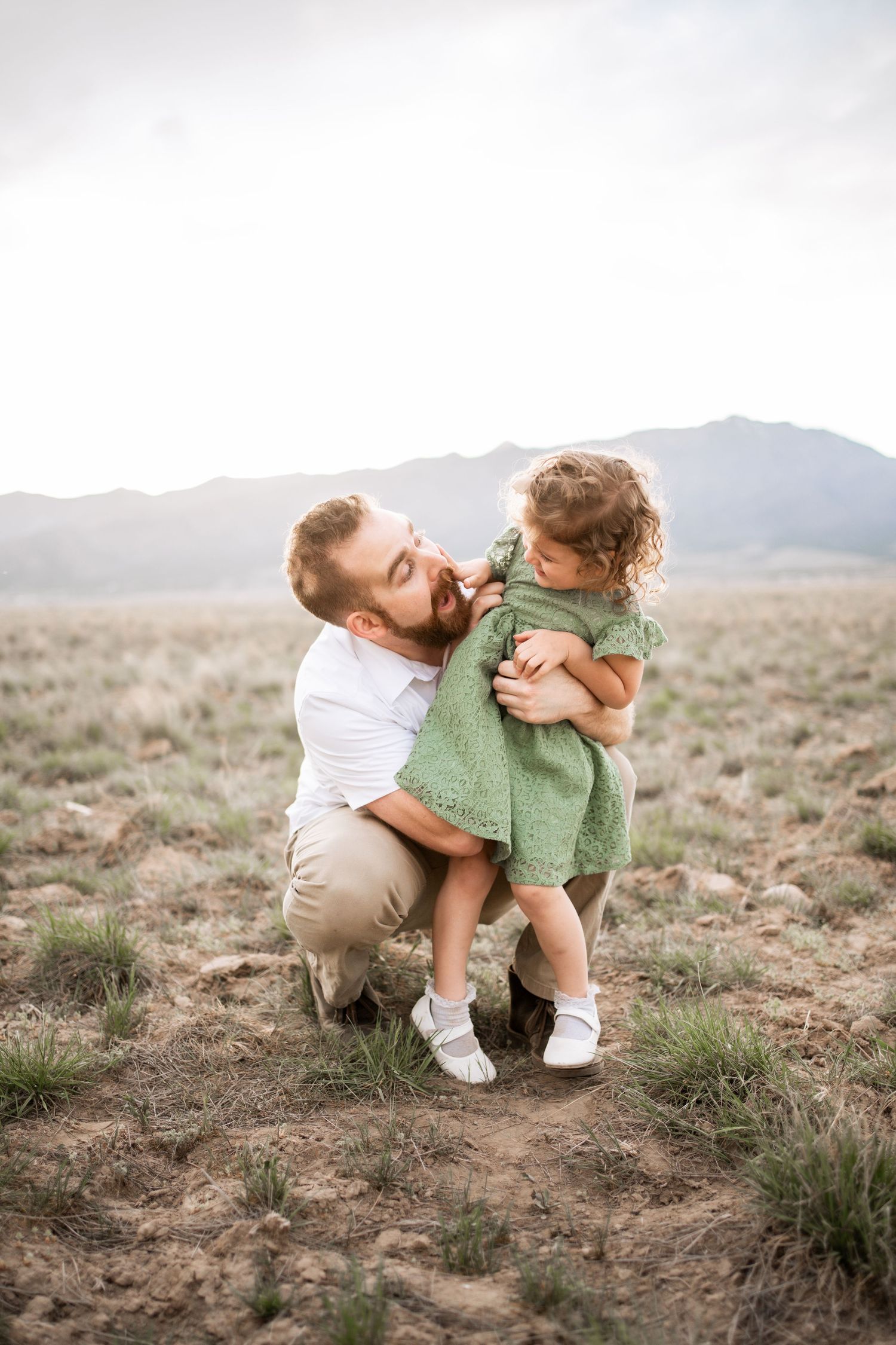 Father kneeling in field holding young daughter in green dress with mountains in background.