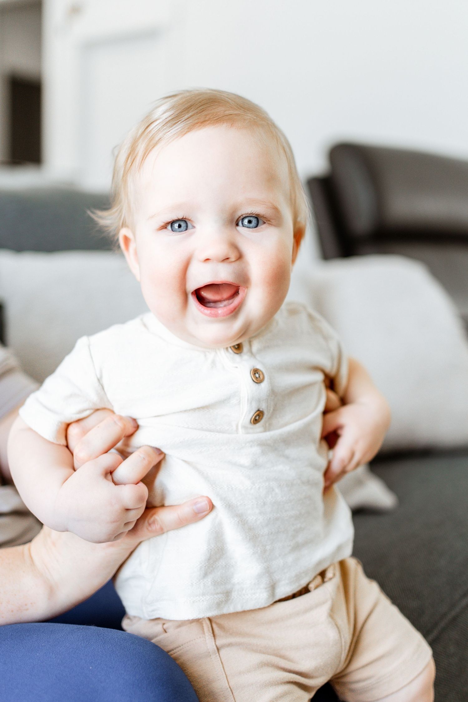 A happy baby wearing a white henley shirt and beige pants sits on a couch while being held by adult hands.