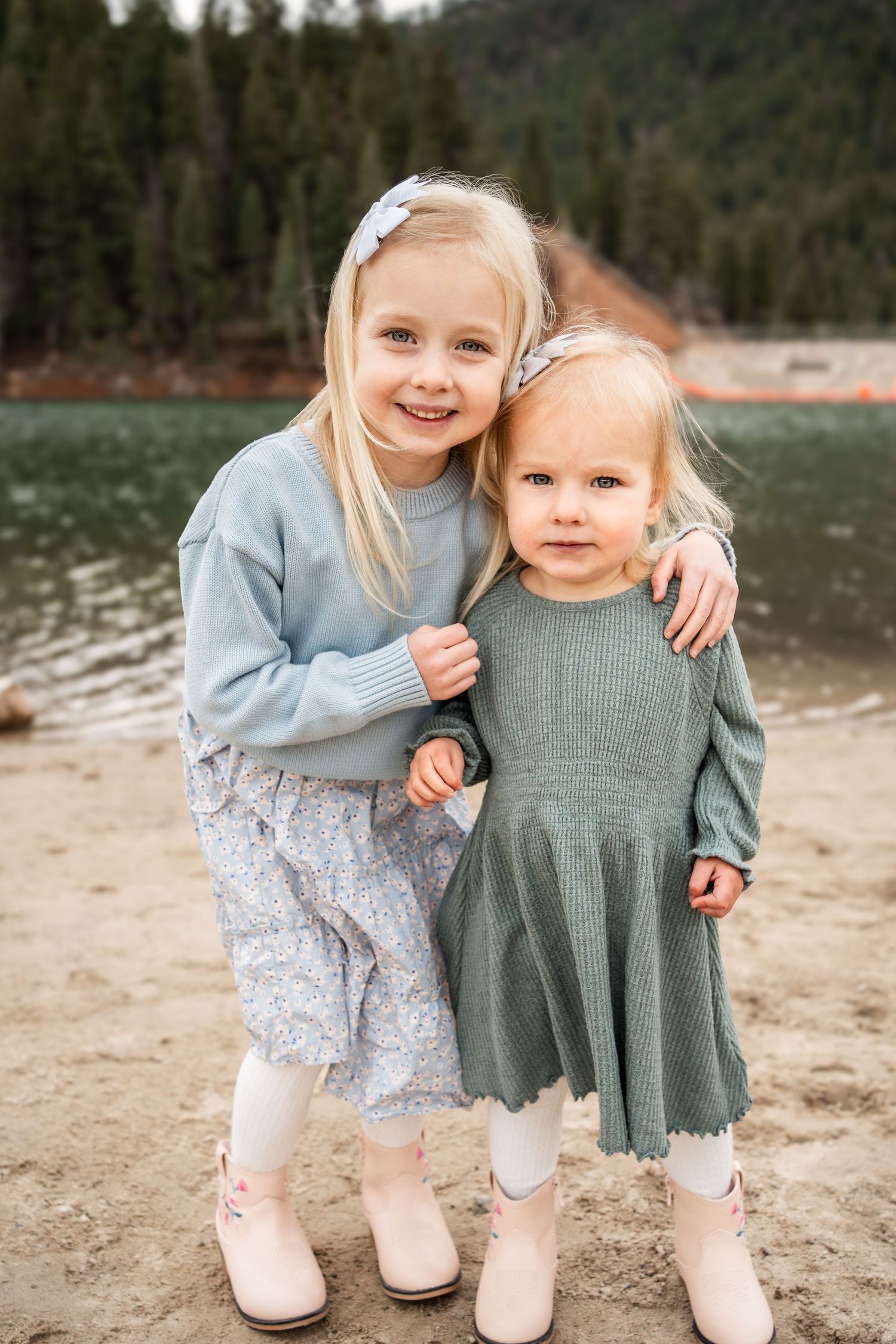Two children in light blue and sage green dresses stand together on a sandy beach with evergreen trees in background.