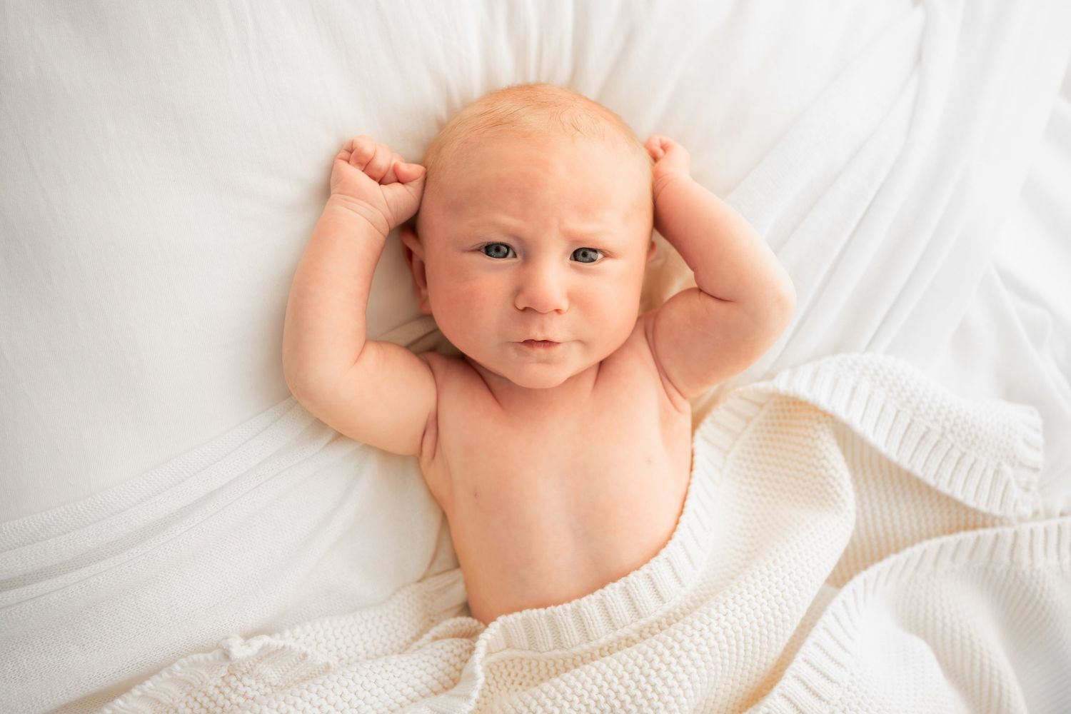 Newborn baby lying on a soft white blanket with both arms raised beside the head, looking calmly at the camera.