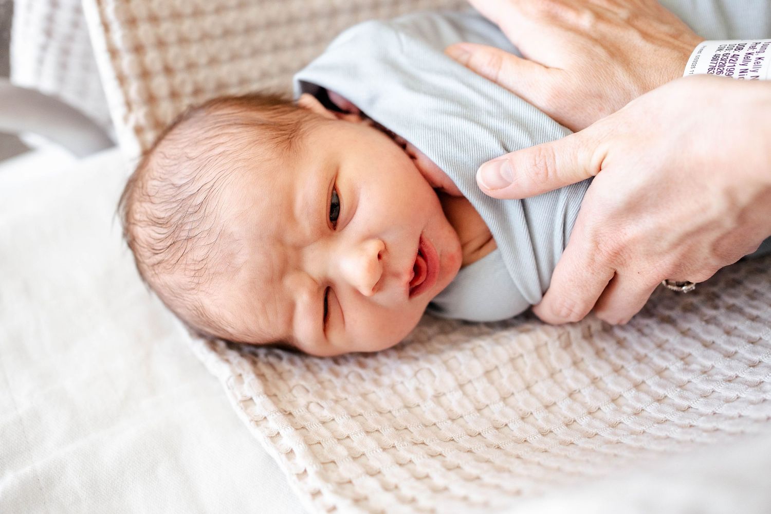Newborn baby swaddled in light blue blanket being held by adult hands in hospital setting.