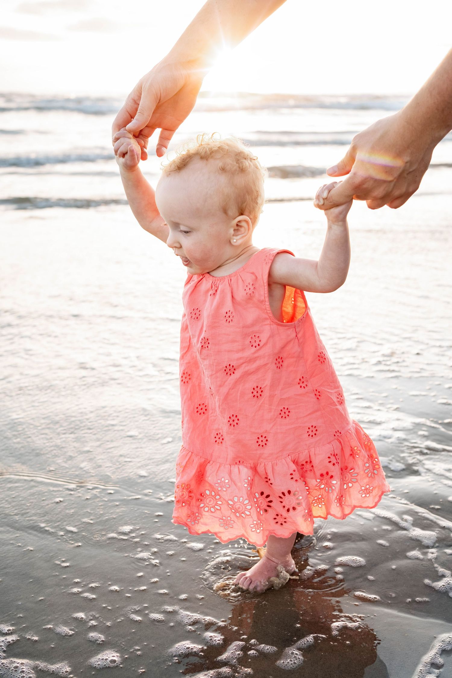 A baby girl in a pink dress takes her first steps on a sandy beach at sunset, guided by adult hands.