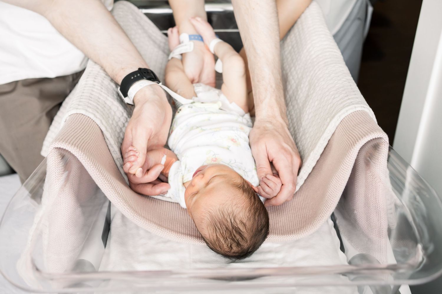 Newborn baby lying in hospital bassinet being gently held by parent hands after birth.
