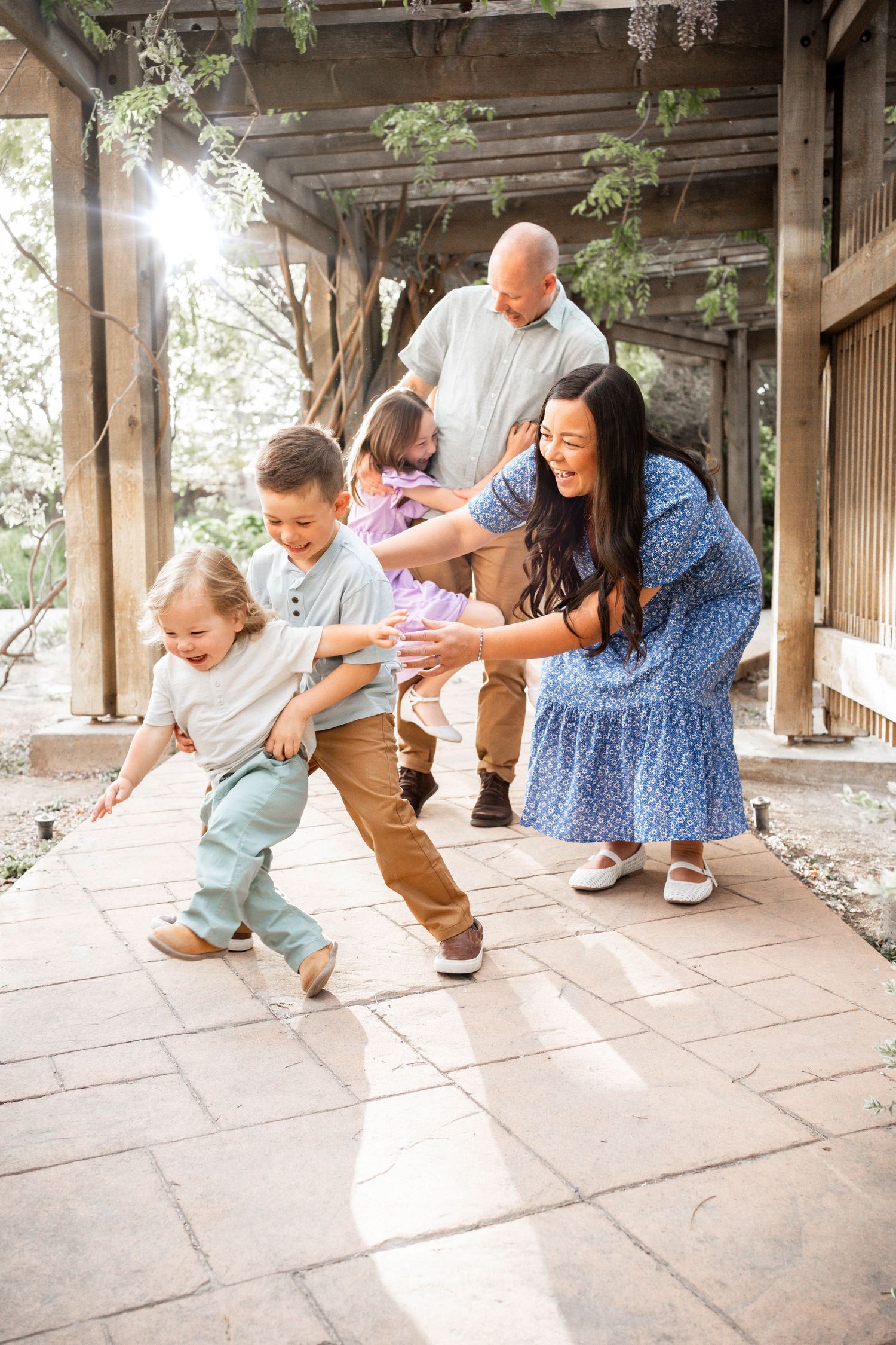 Smiling family laughing and playing on a stone patio under a wooden pergola as two young boys run toward a woman in a blue dress.
