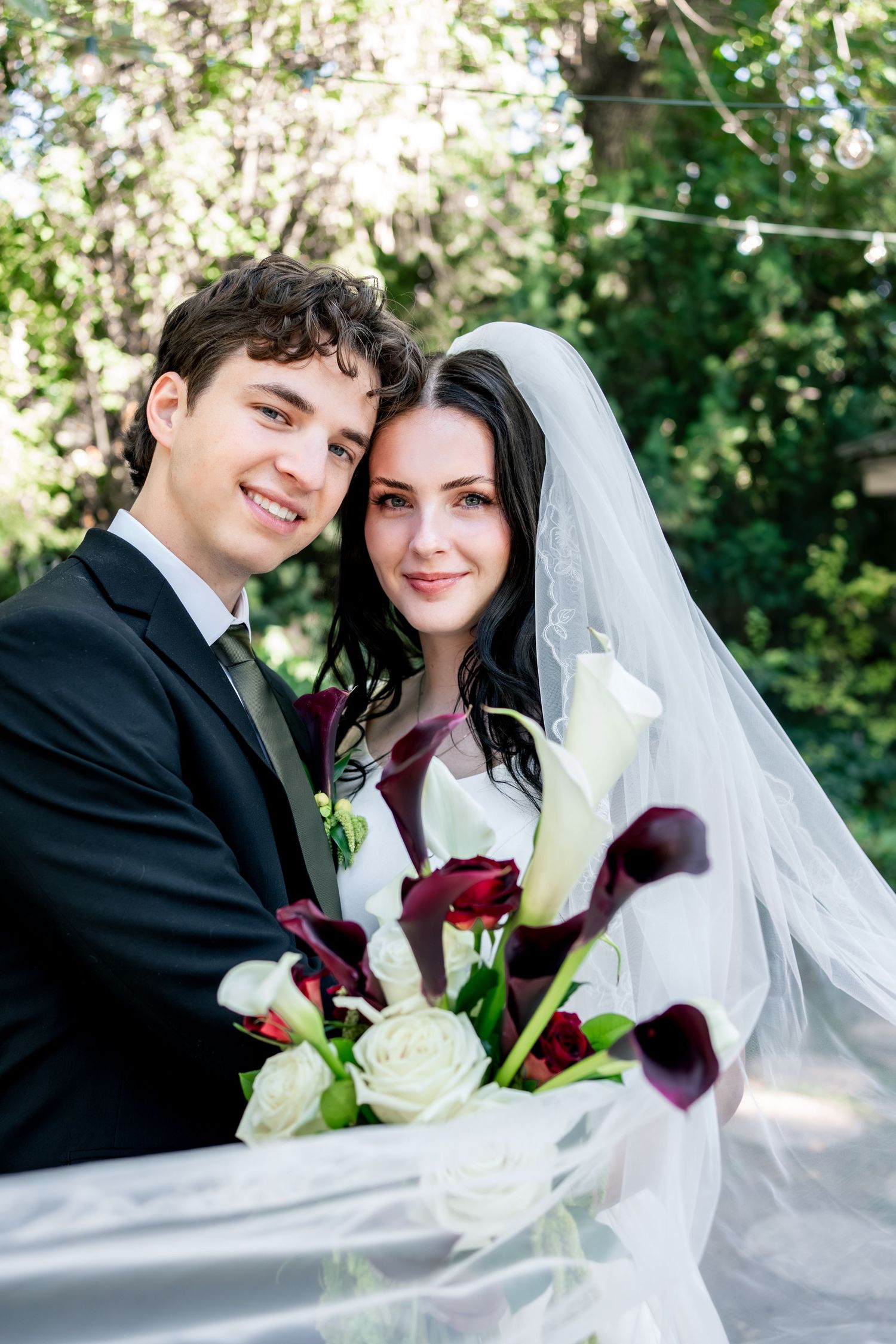 Smiling bride and groom embrace outdoors, holding a bouquet of white roses and dark burgundy calla lilies beneath a flowing veil.