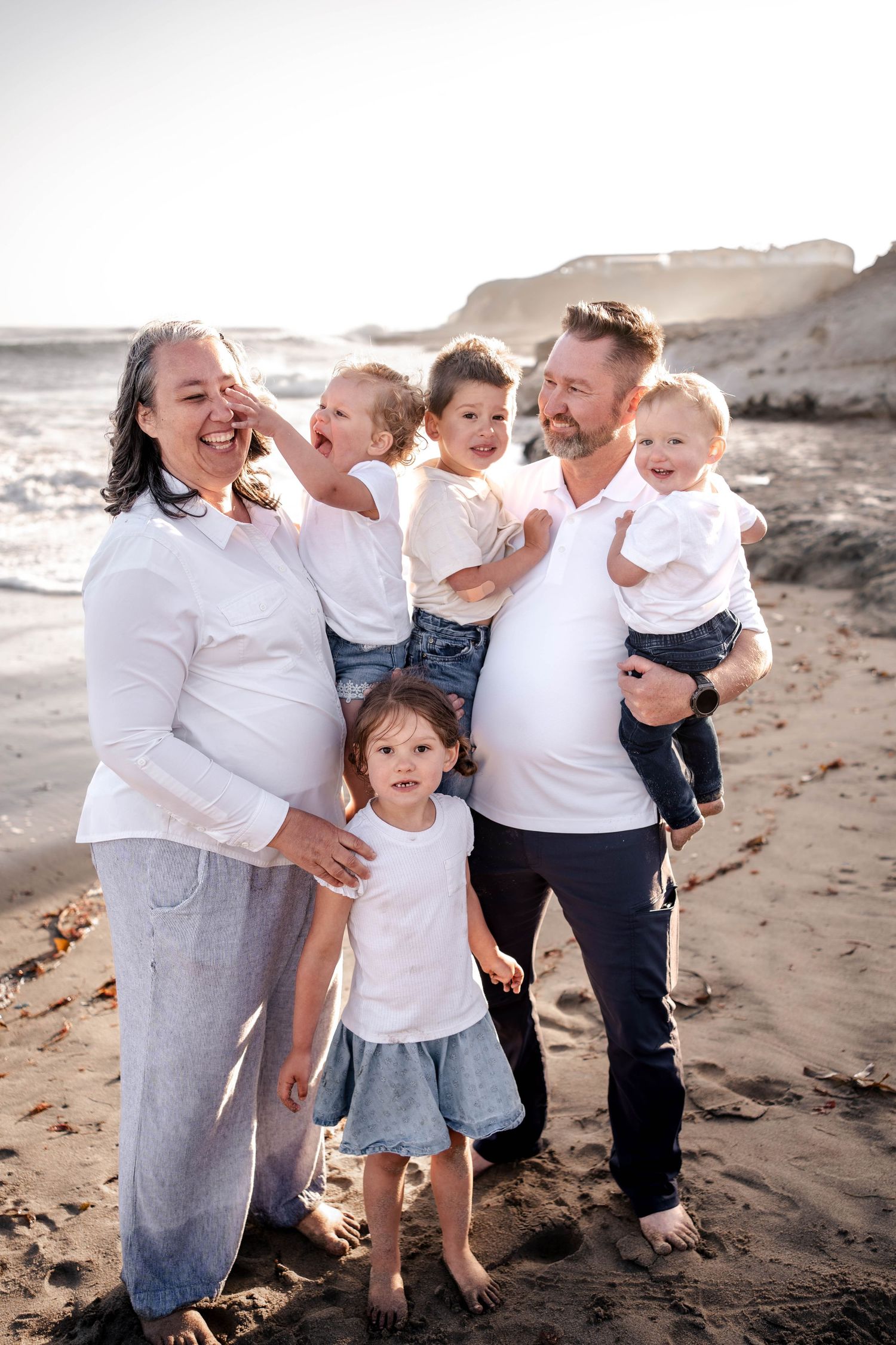 Happy family of six posing together on a sunny beach, parents holding children in white tops and denim.