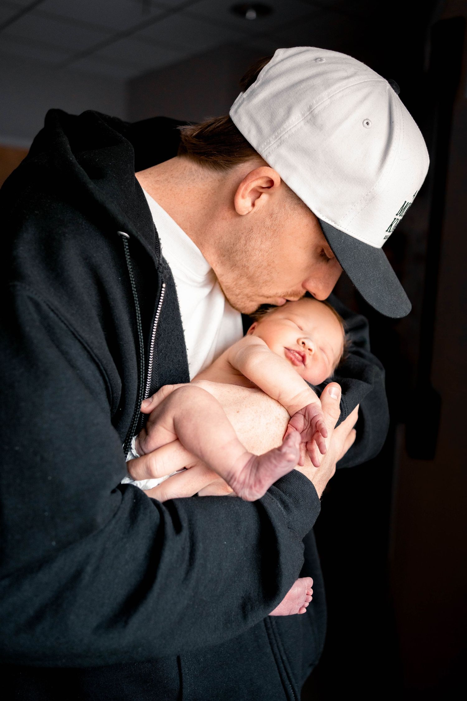 A father in a white cap and black hoodie tenderly kisses his sleeping newborn baby cradled in his hands.