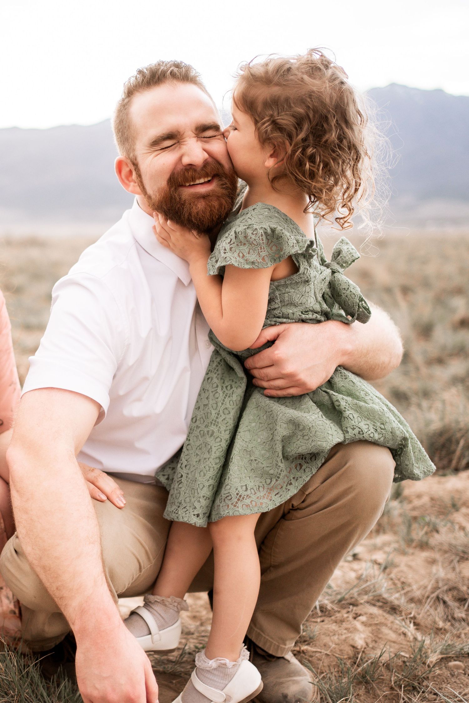 A young girl in a green dress kisses her bearded father on the cheek outdoors with mountains in the background.